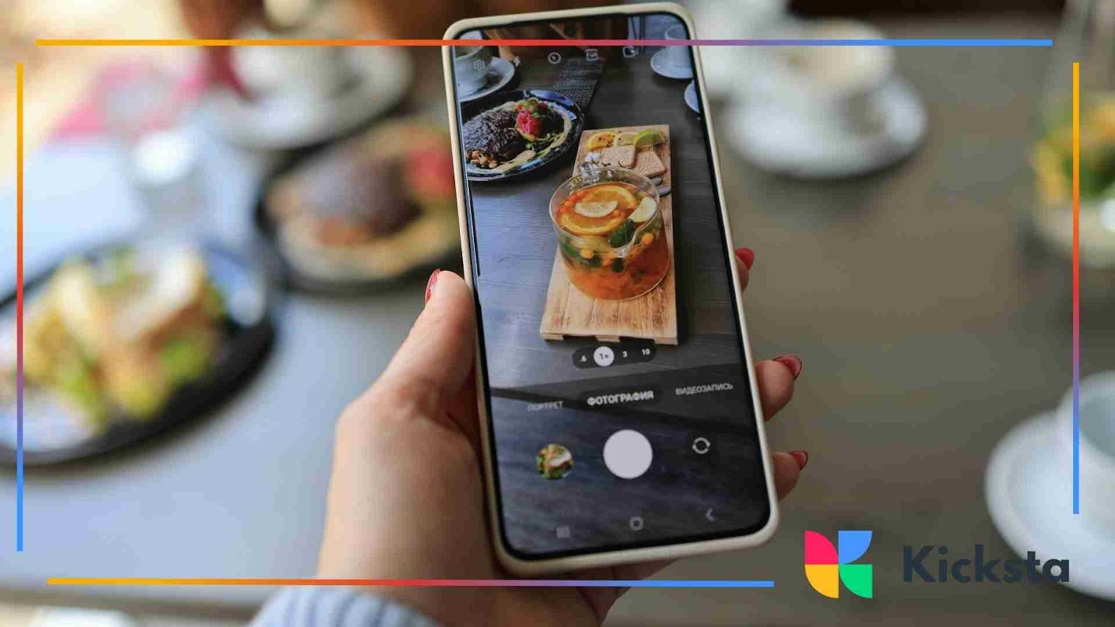 A hand holding a smartphone captures a photo of a colorful food dish on a restaurant table with plates and drinks in the background.