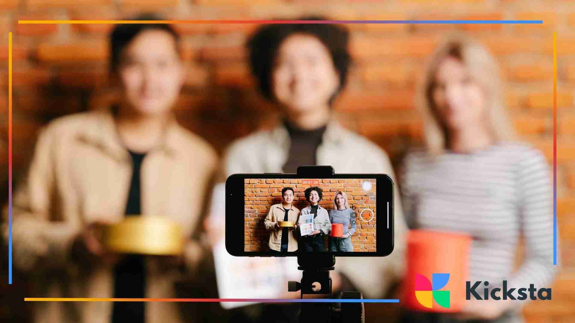 Smartphone on a tripod recording three people standing in front of a brick wall, with the phone screen showing them clearly while the background remains blurred.