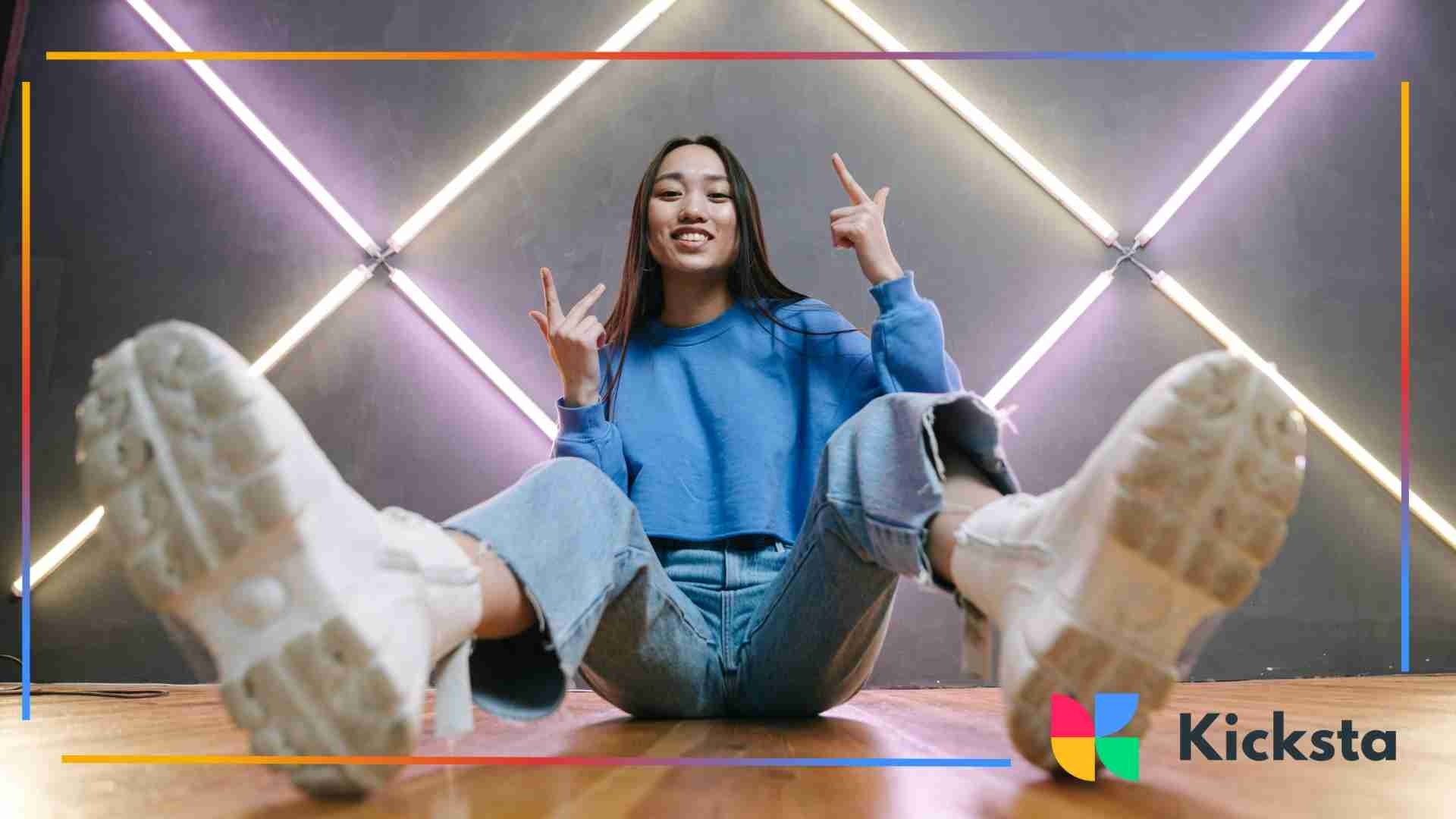 Young woman in casual jeans and a blue sweatshirt sitting on the floor posing confidently with bright diagonal neon lights behind her.