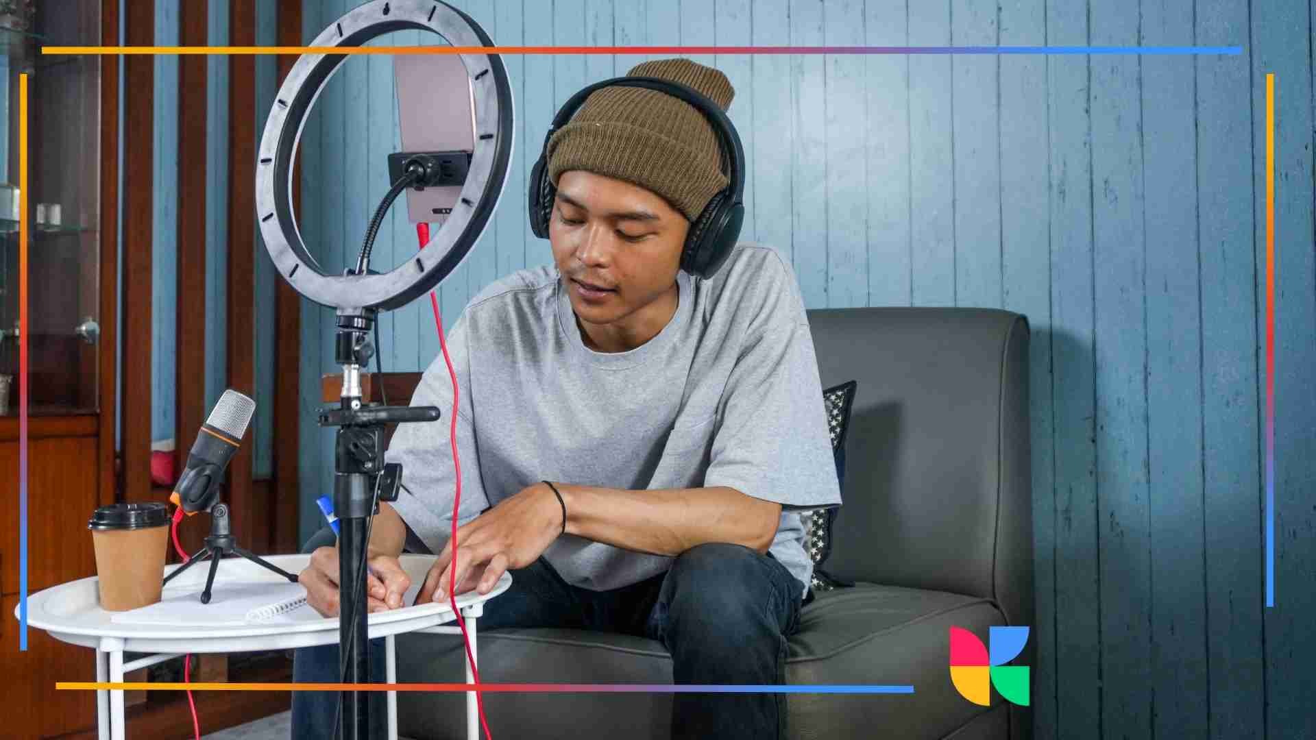 A content creator wearing headphones writes notes while recording with a smartphone mounted on a ring light and a microphone on a small table.