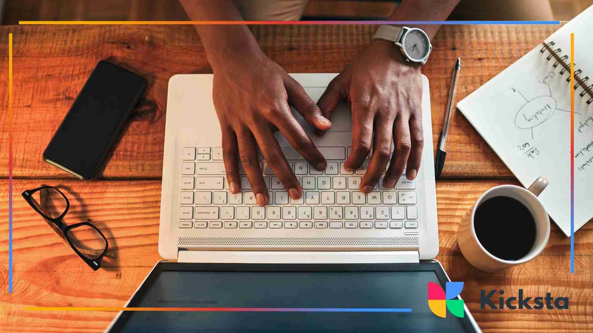 A person typing on a laptop at a wooden desk with a notebook, coffee cup, smartphone, pen, and glasses placed around them.