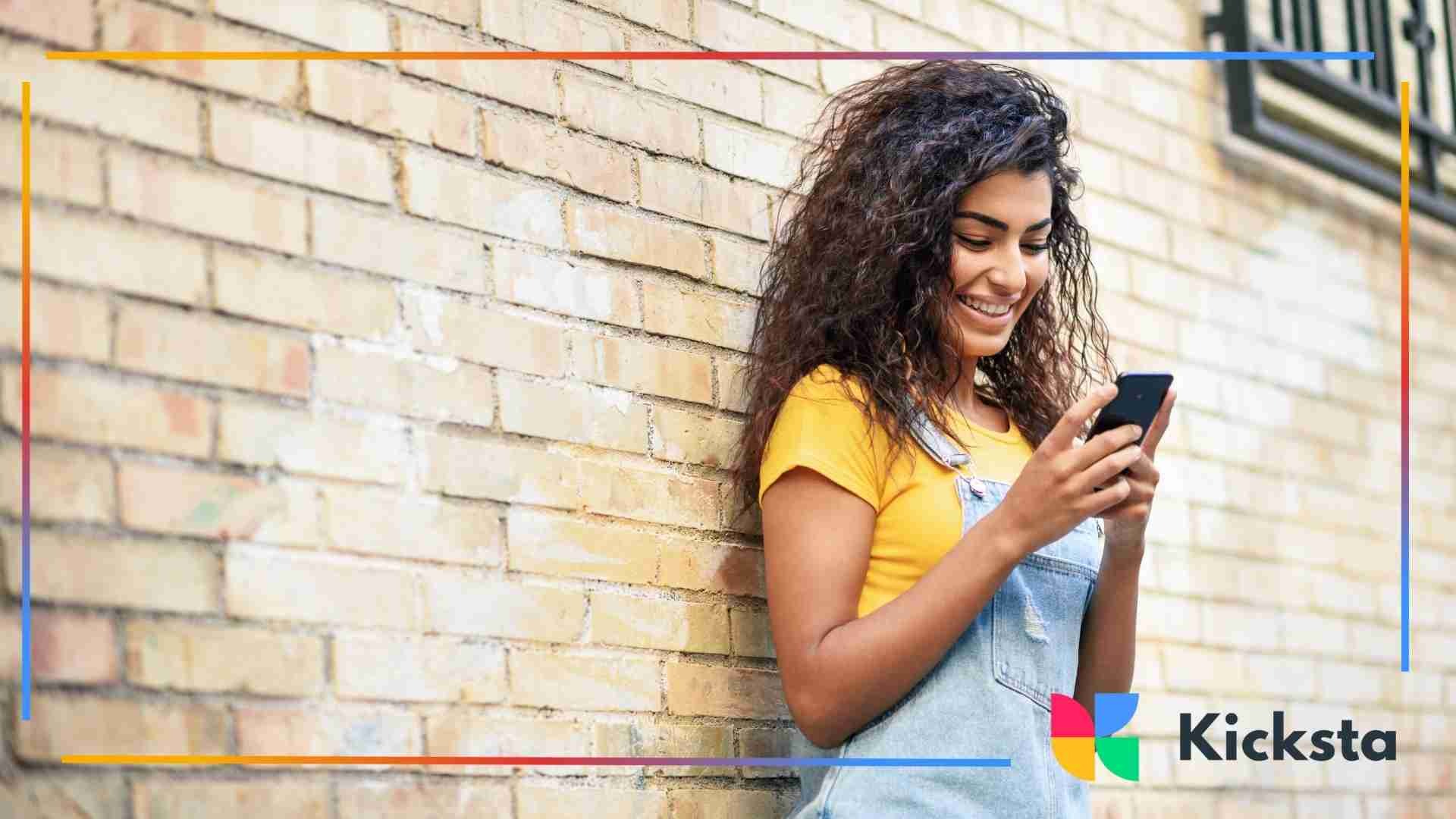 A young woman standing by a brick wall smiling as she looks down at her phone.