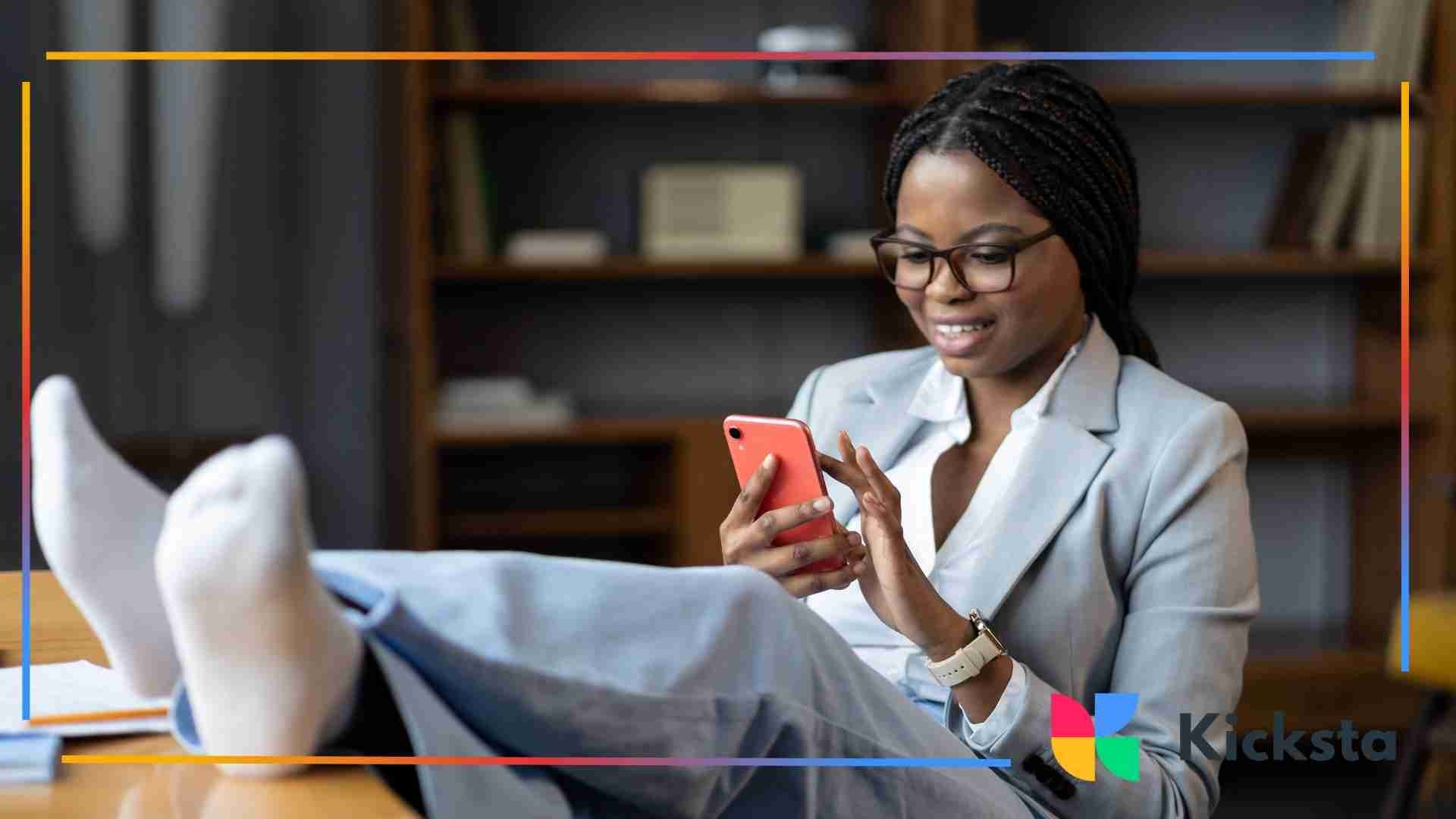 A woman in business attire relaxing with her feet up on a desk while smiling at her phone.