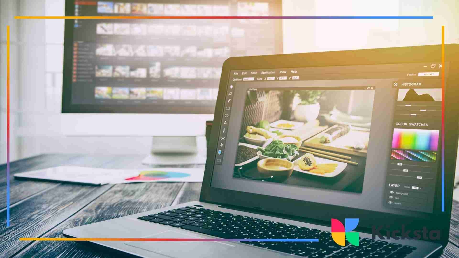 A laptop displaying a photo editing program with food images on the screen, sitting on a wooden desk with a large desktop monitor blurred in the background.