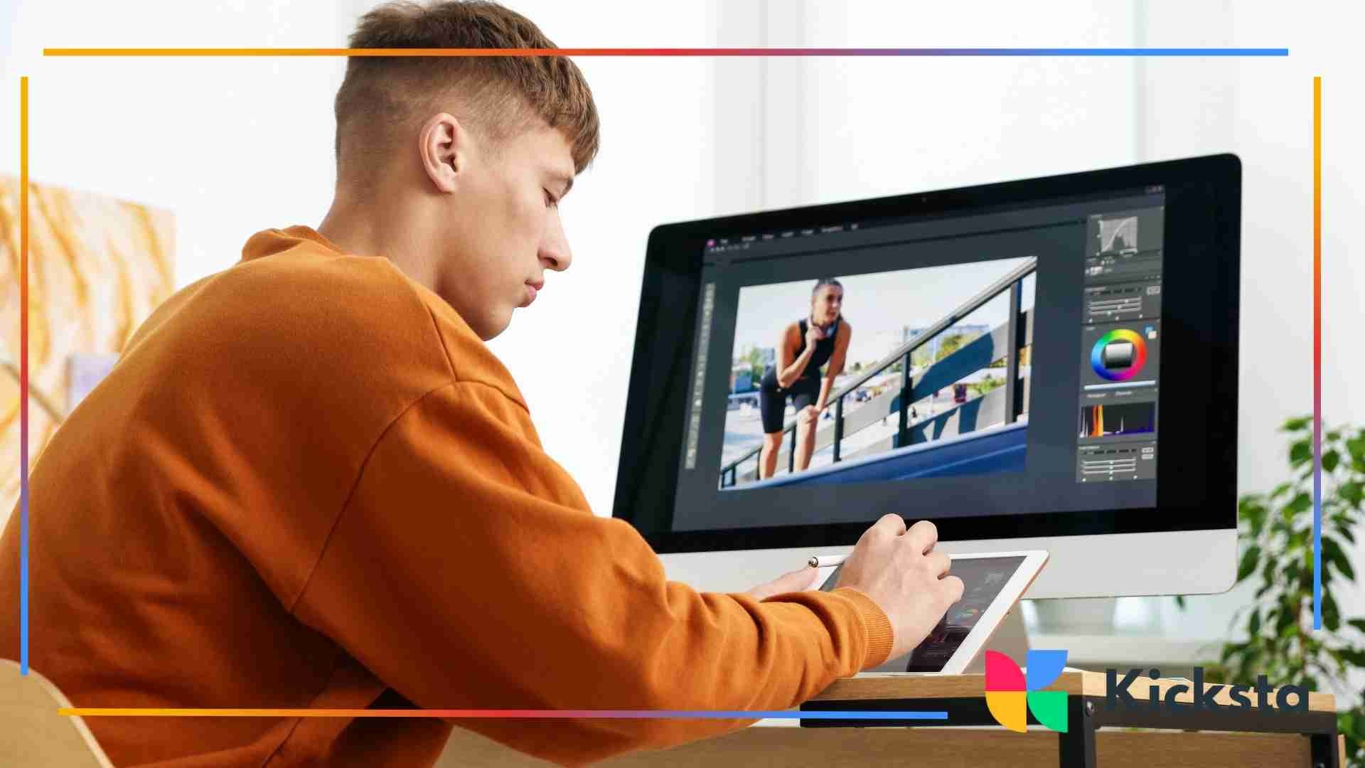 A young man using a tablet while editing a photo on a large desktop monitor, working in a bright creative workspace.