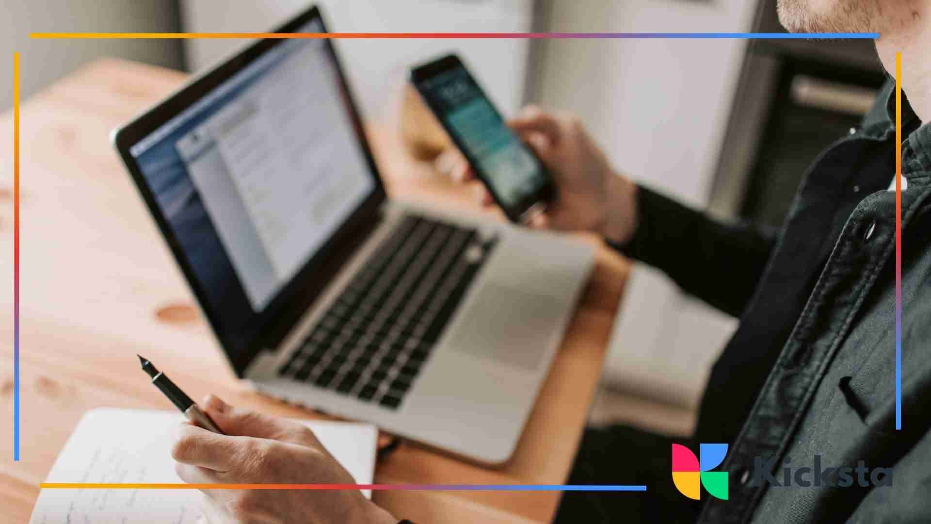A person working at a desk while holding a smartphone, using a laptop, and writing notes in a notebook.