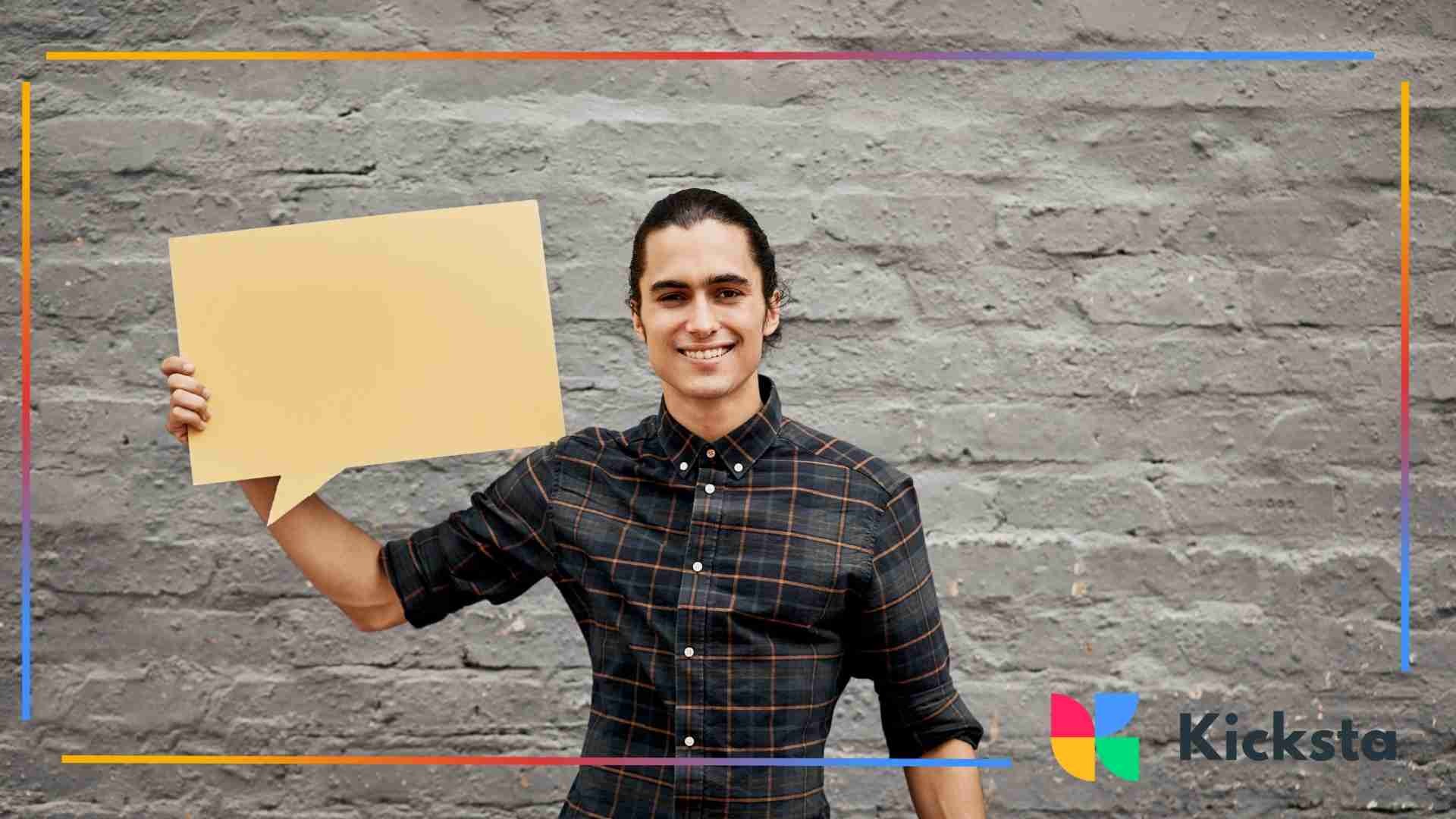 Smiling man standing against a brick wall holding a large blank speech-bubble sign.