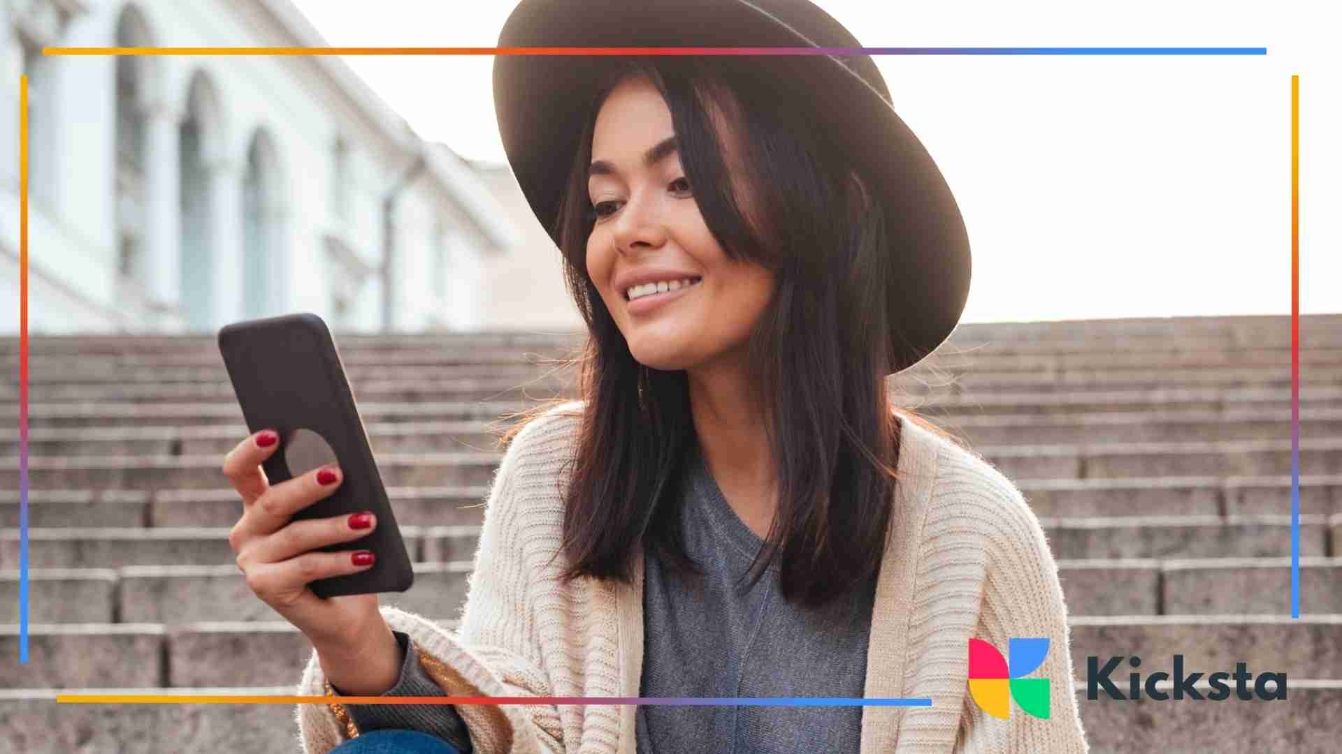 Woman wearing a black hat and casual outfit sitting on outdoor steps and smiling at her phone.
