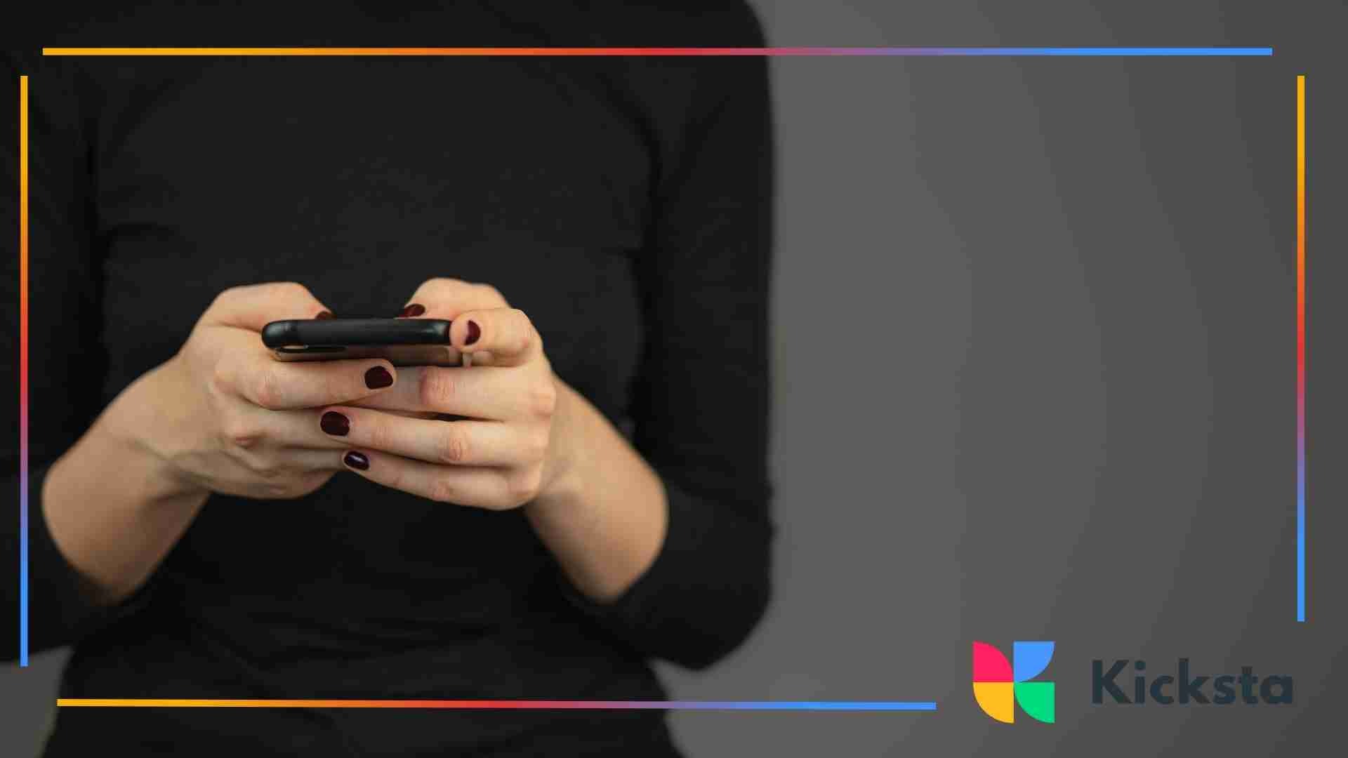 Person dressed in a black top holding a smartphone with both hands against a dark background.