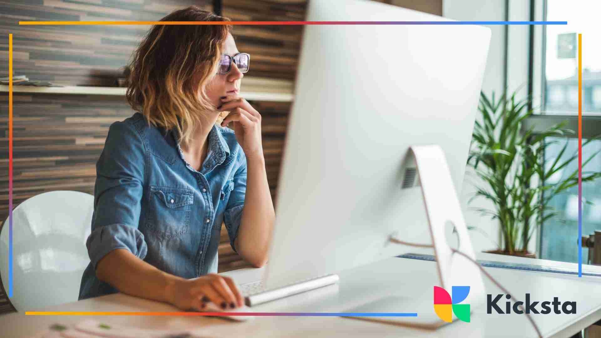 A woman wearing glasses sitting at a desk, thinking while working on a large desktop computer.