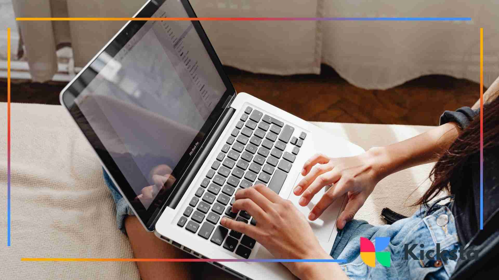 Hands typing on a laptop while sitting on a couch in a relaxed home setting.