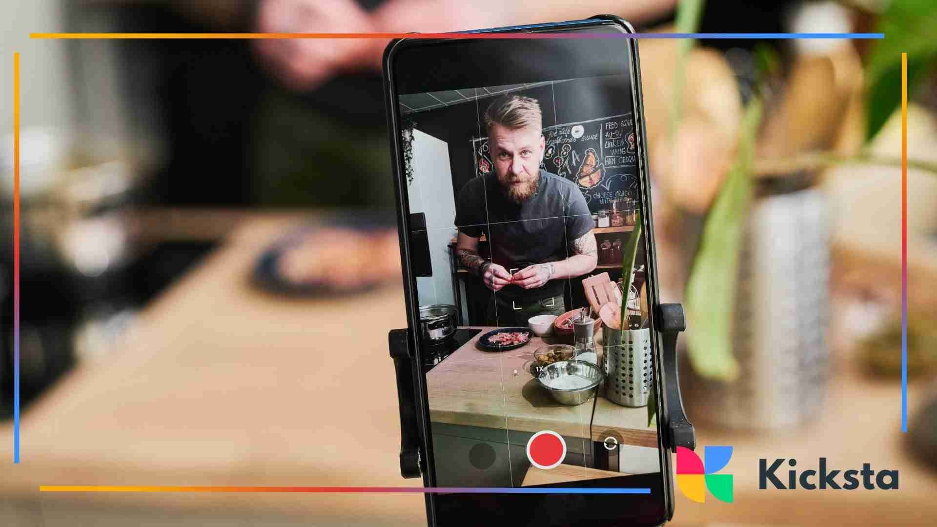 A smartphone recording a bearded chef preparing food in a kitchen, with cooking tools and ingredients visible around him.