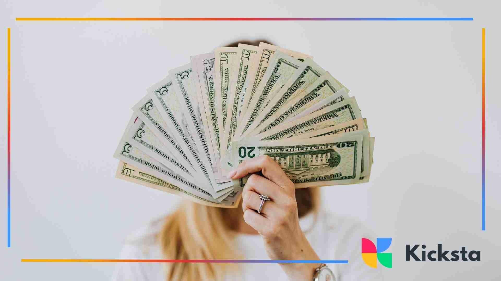 A person holding a large fanned-out stack of U.S. dollar bills in front of their face, showing various denominations.