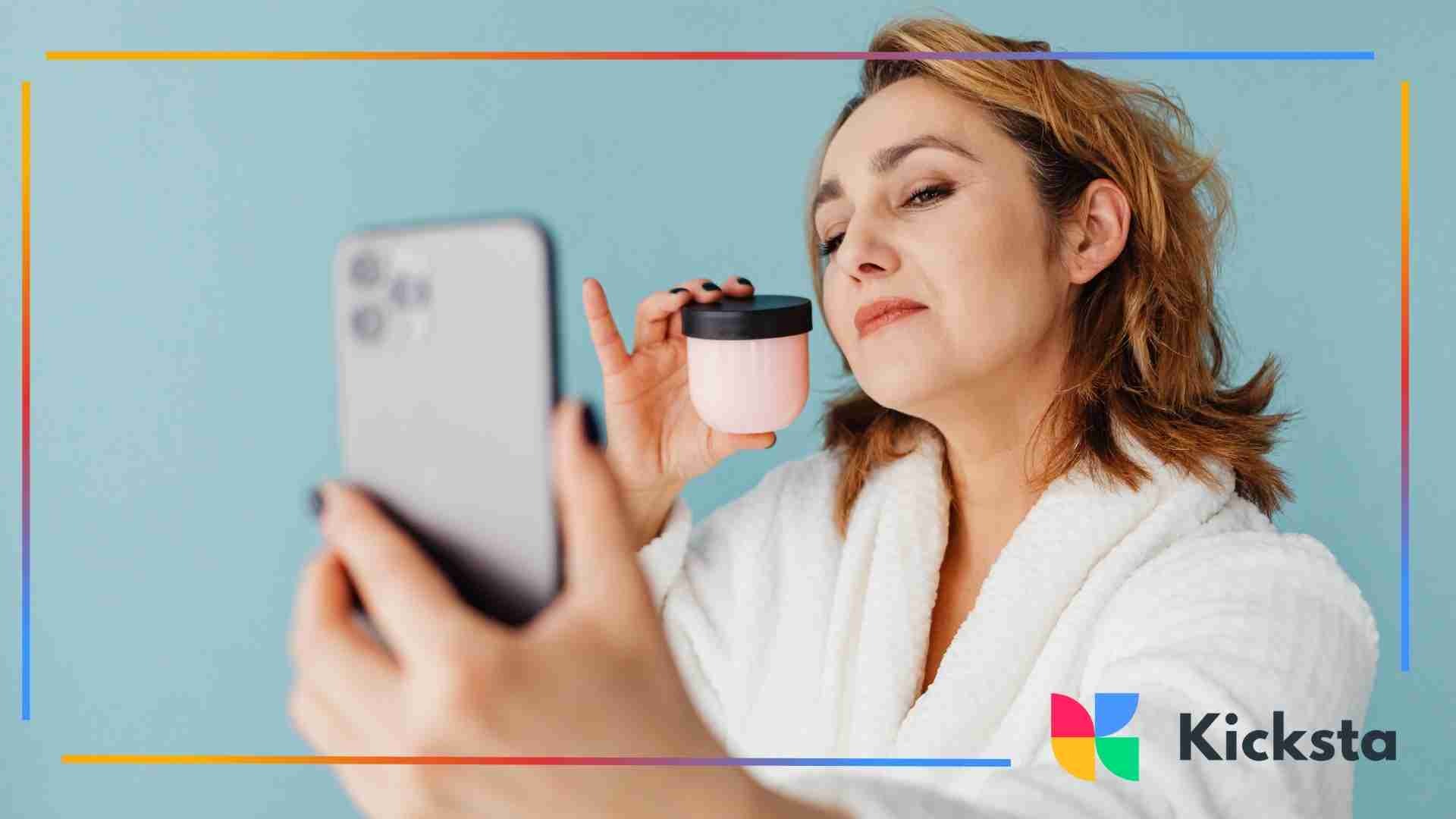 A woman wearing a white robe holding a pink cosmetic jar and taking a selfie against a blue background.