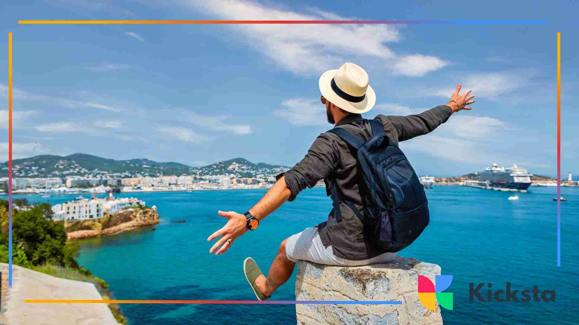 Man with backpack sitting on a stone ledge overlooking a blue ocean and coastal city with arms outstretched.