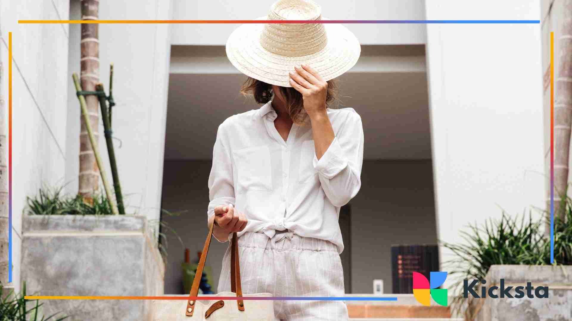 Woman wearing a white outfit and straw hat standing outdoors, holding a handbag.
