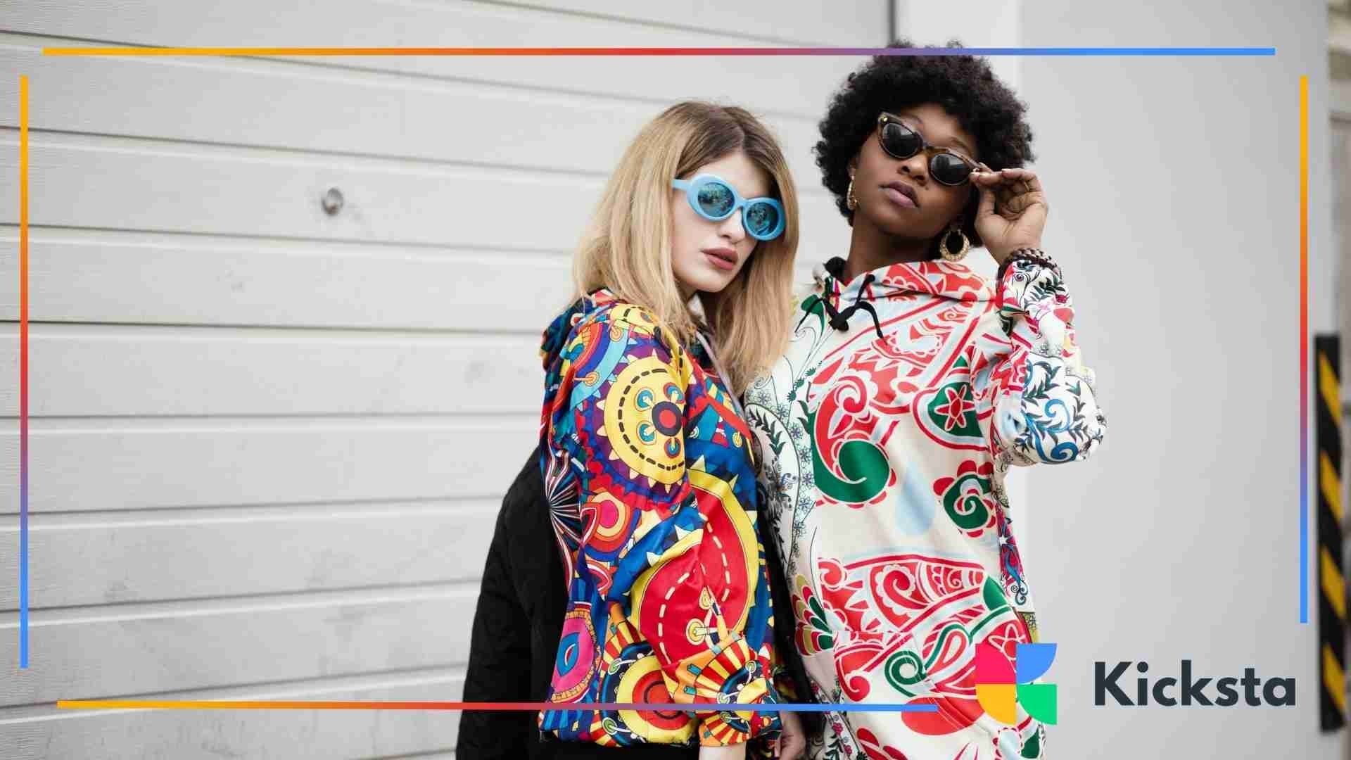 Two stylish women wearing colorful patterned hoodies and sunglasses posing against a light wall.
