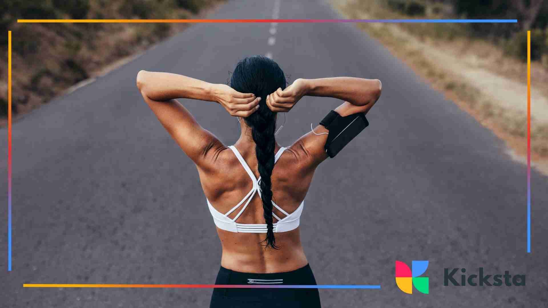 Fit woman stretching her arms on an empty road before a workout, wearing a sports bra and armband.