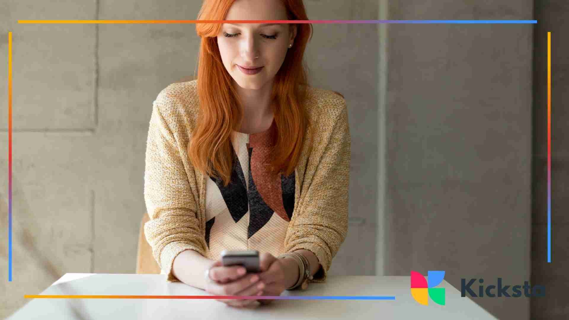 Woman with red hair sitting at a table and looking down at her smartphone in a modern indoor space.