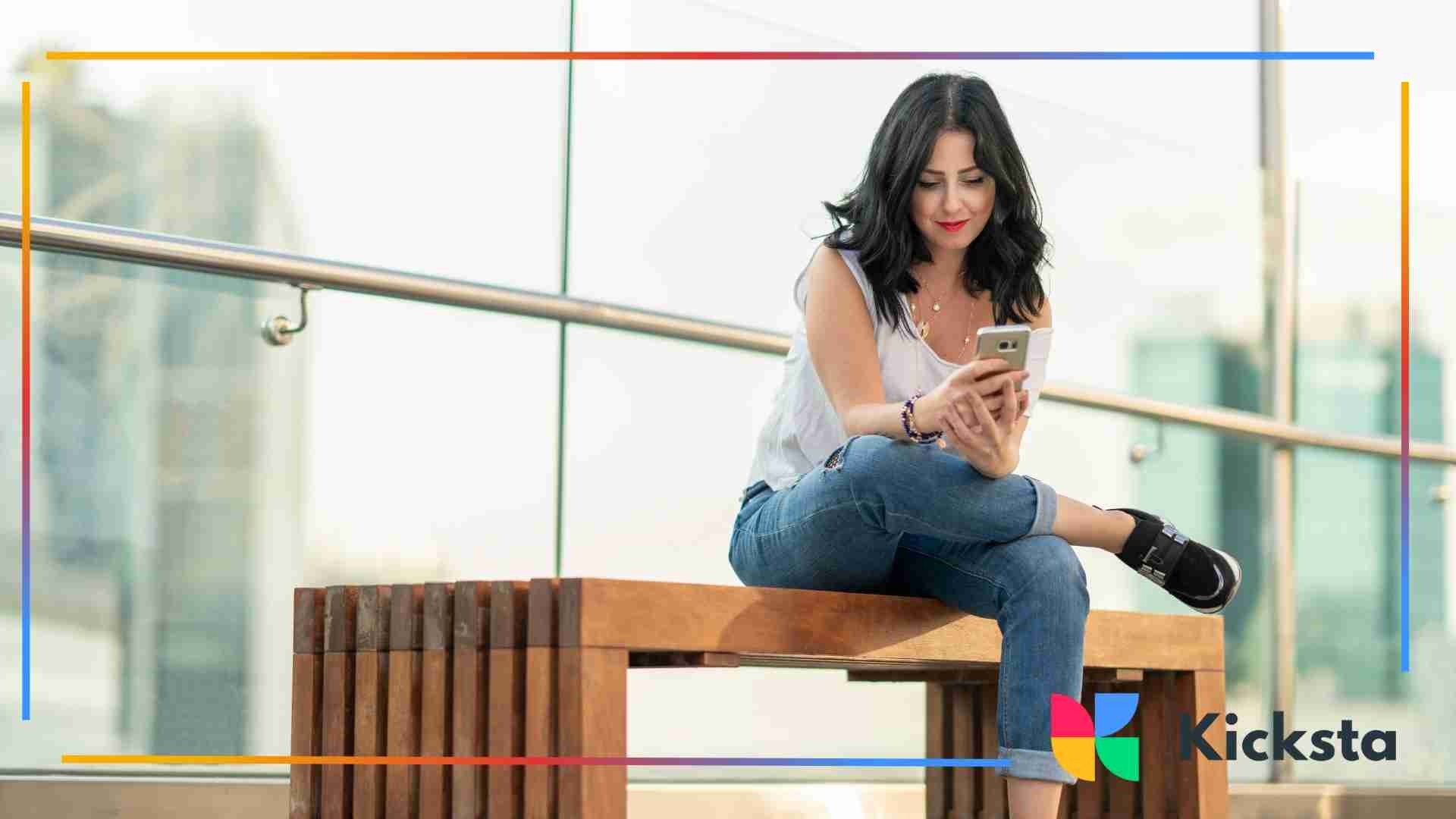 Woman sitting cross-legged on a wooden bench, smiling while using her smartphone in an outdoor setting.