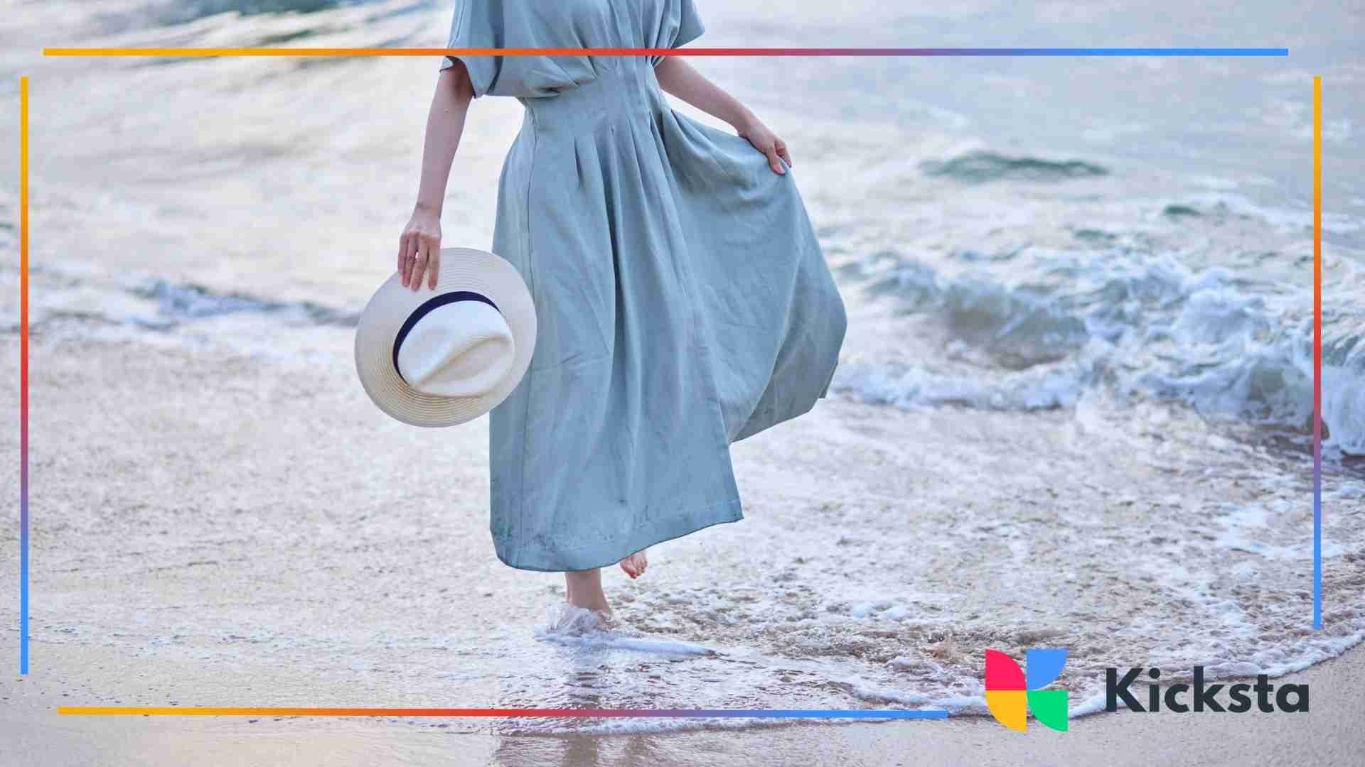 Person in a flowing blue dress walking barefoot along the shoreline while holding a sun hat.