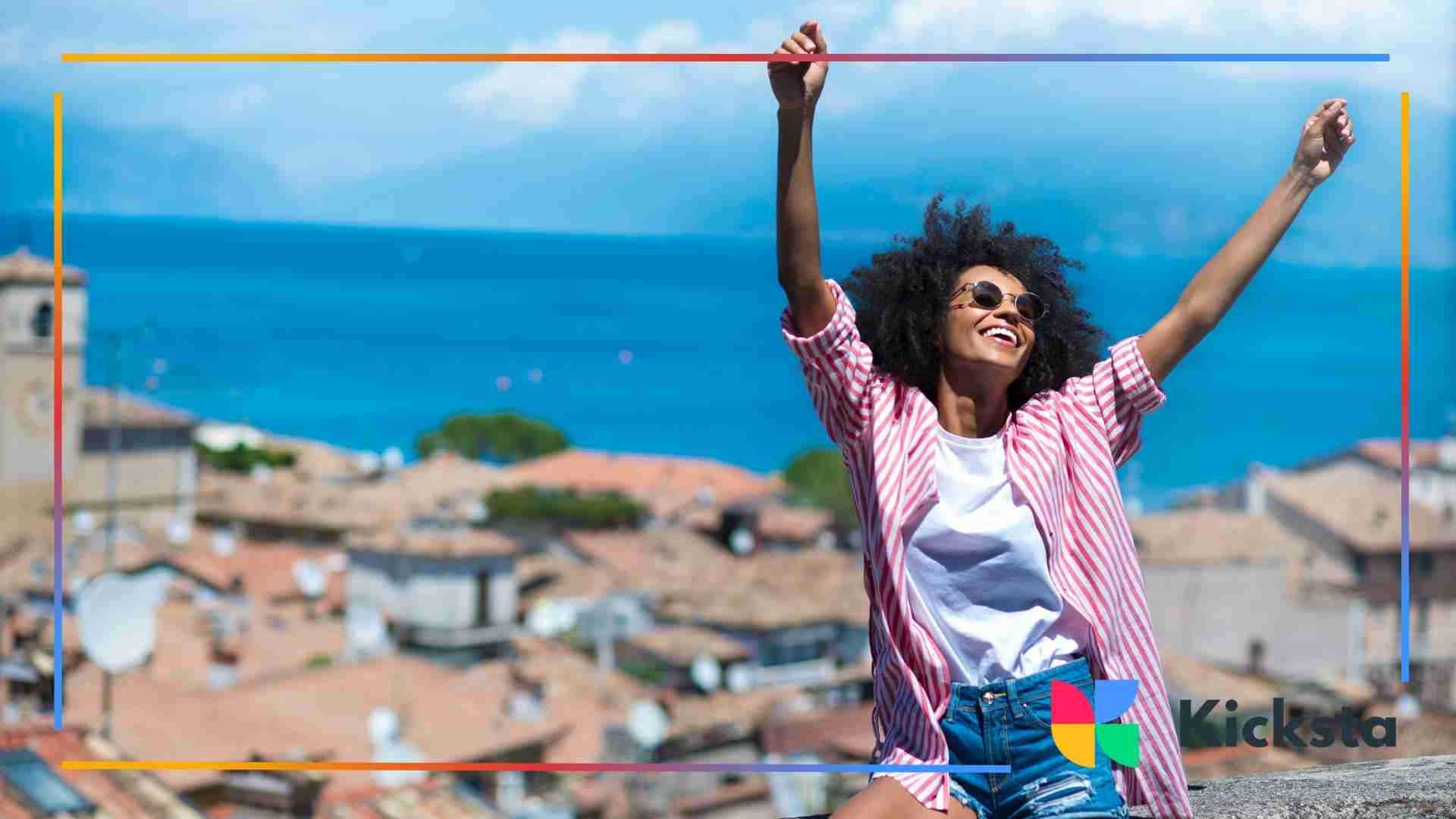 A woman with curly hair raising her arms joyfully in front of a scenic view with rooftops and blue skies.
