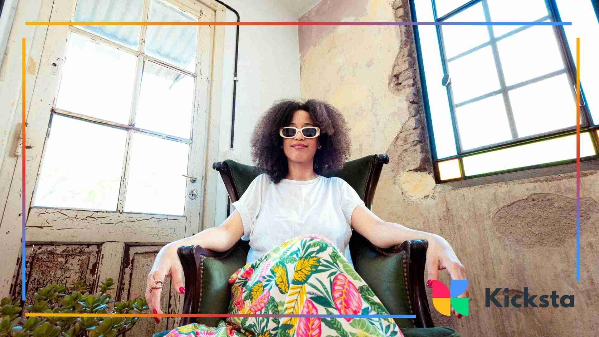 A woman sitting in a vintage chair, wearing sunglasses and a casual white t-shirt, with a plant in the foreground and natural light streaming in through a window.
