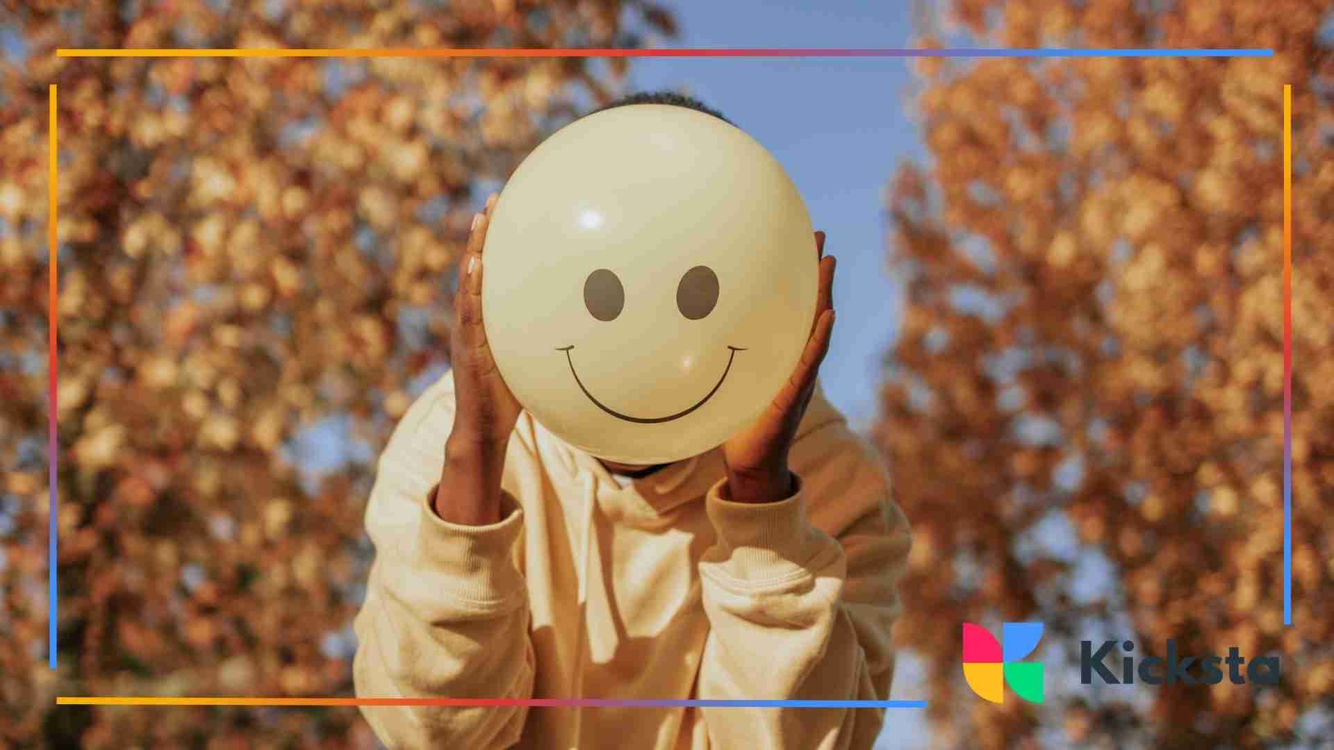 A person holding a yellow balloon with a smiley face in front of a blurred background with golden autumn leaves.