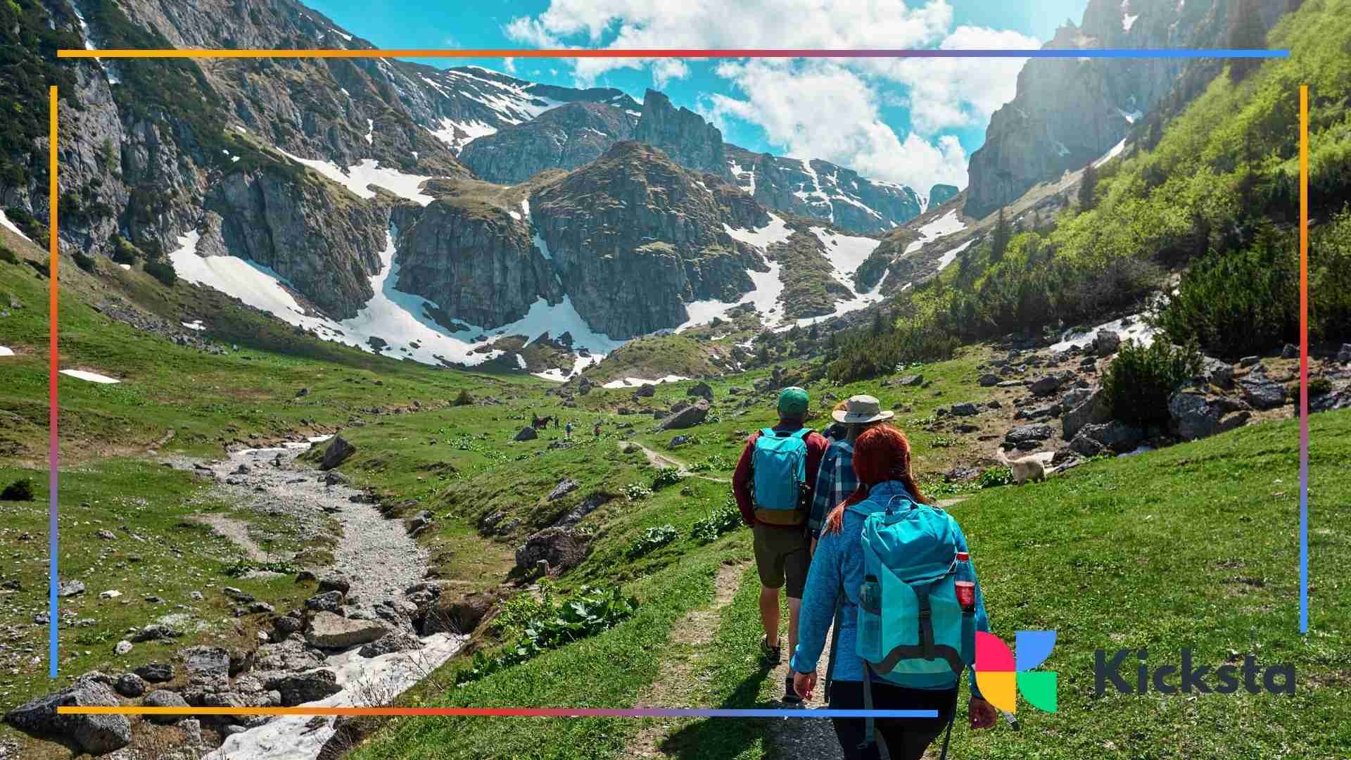 A group of hikers walking along a trail through a green mountain valley with rocky peaks and patches of snow.