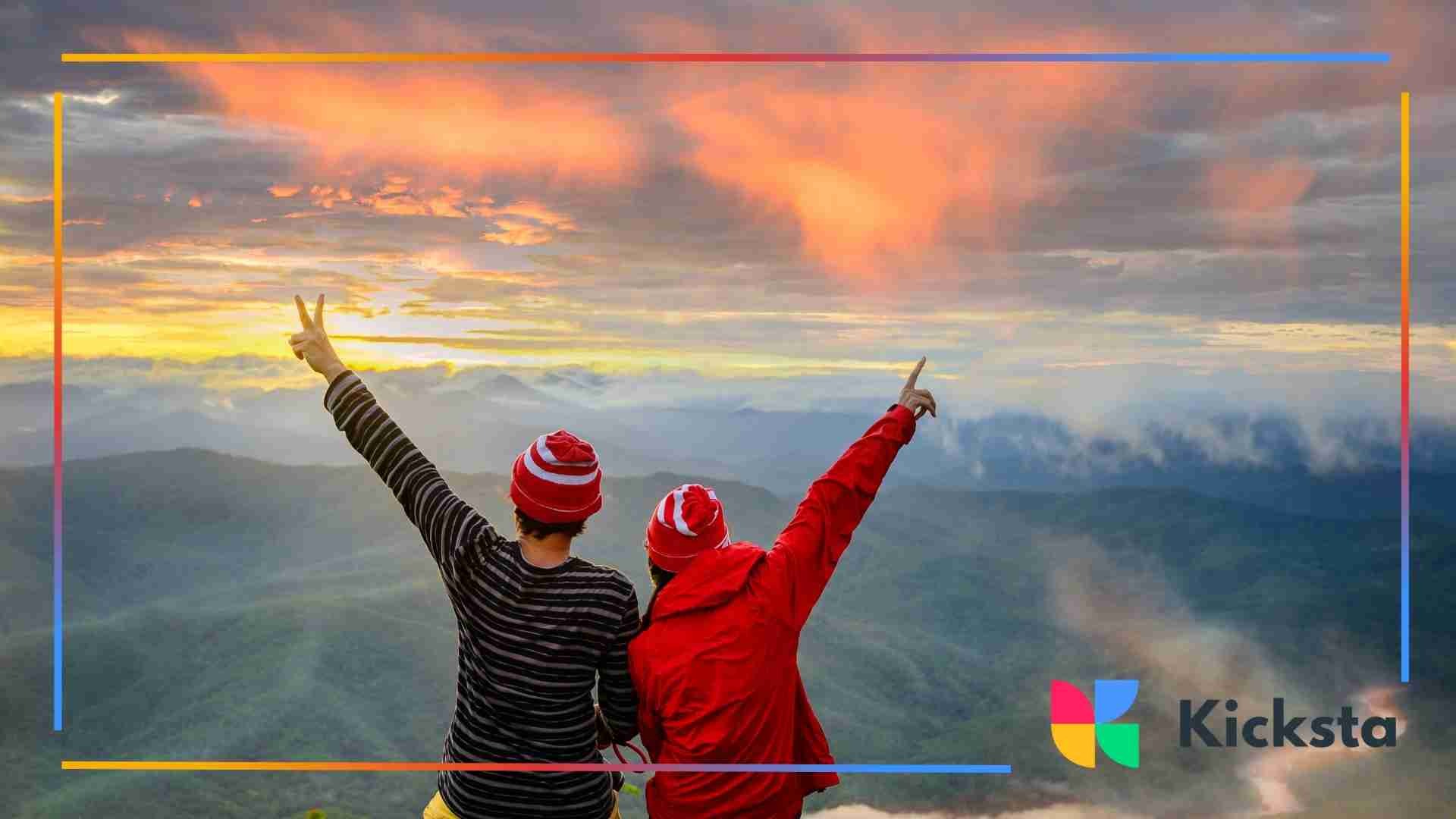 Two people standing on a mountain overlook, raising their arms as the sun sets over layered hills and clouds.