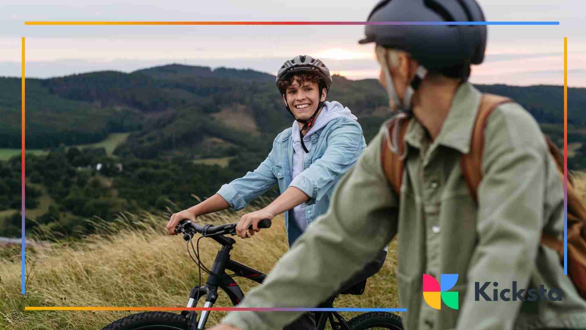 Two cyclists wearing helmets pause on a grassy hillside, smiling with rolling green hills in the background.