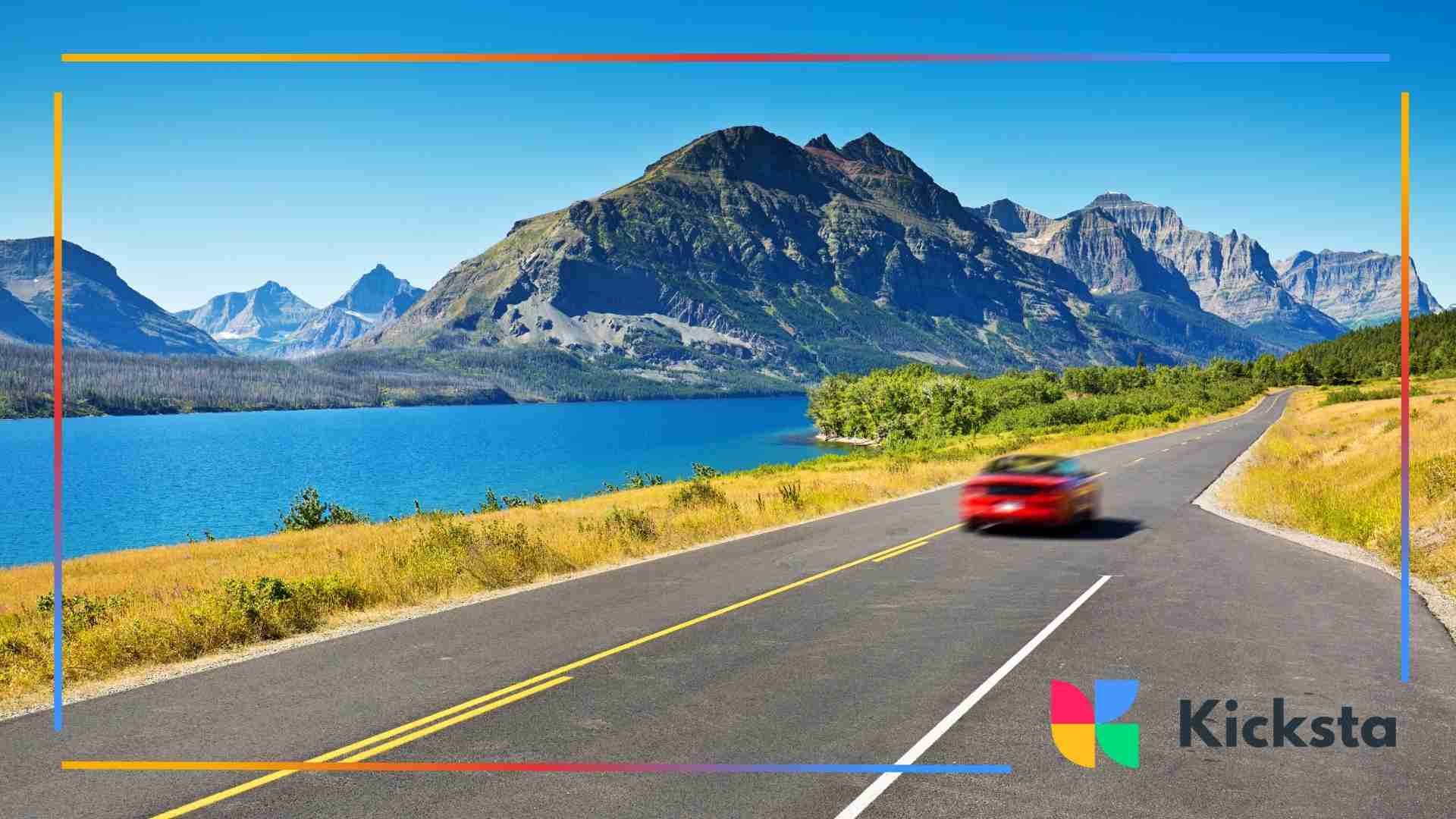 A winding road beside a bright blue lake with rugged mountains rising under a clear sky.