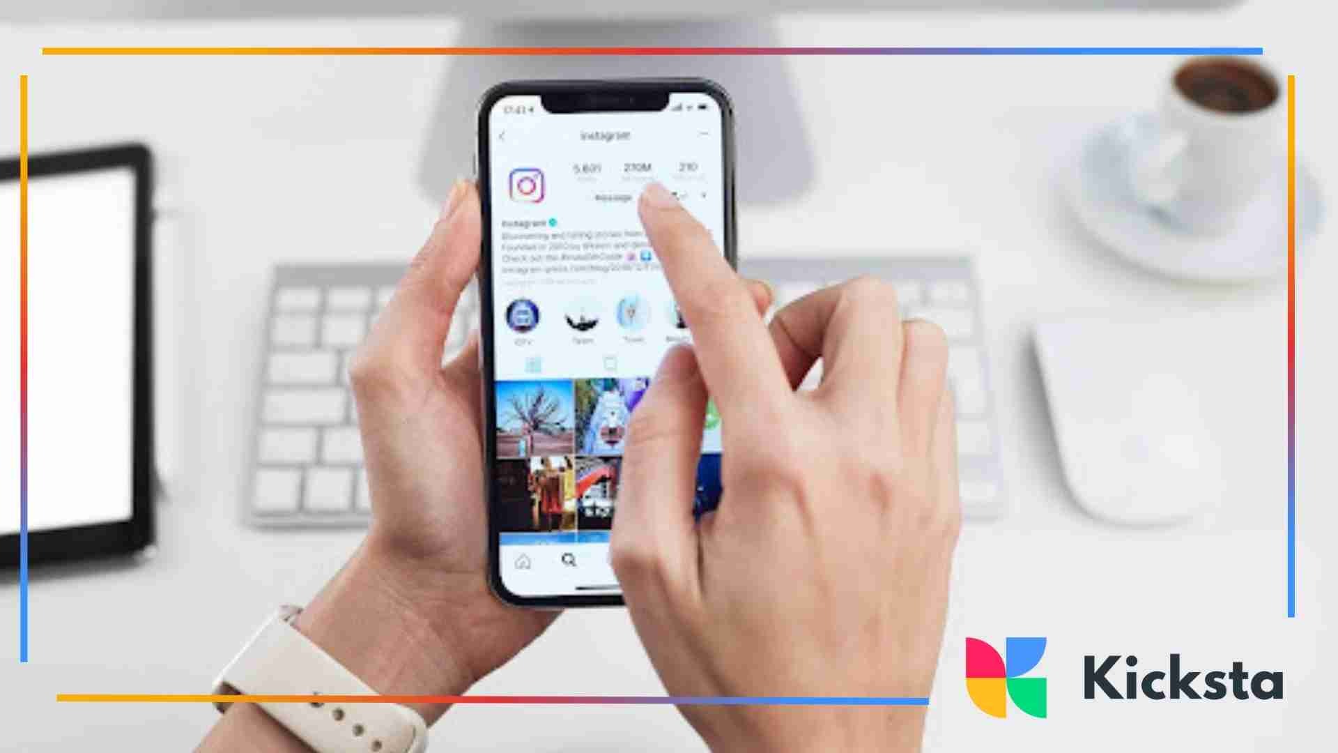 Person using a smartphone to view an Instagram profile on a desk with a keyboard, mouse, and coffee cup nearby.