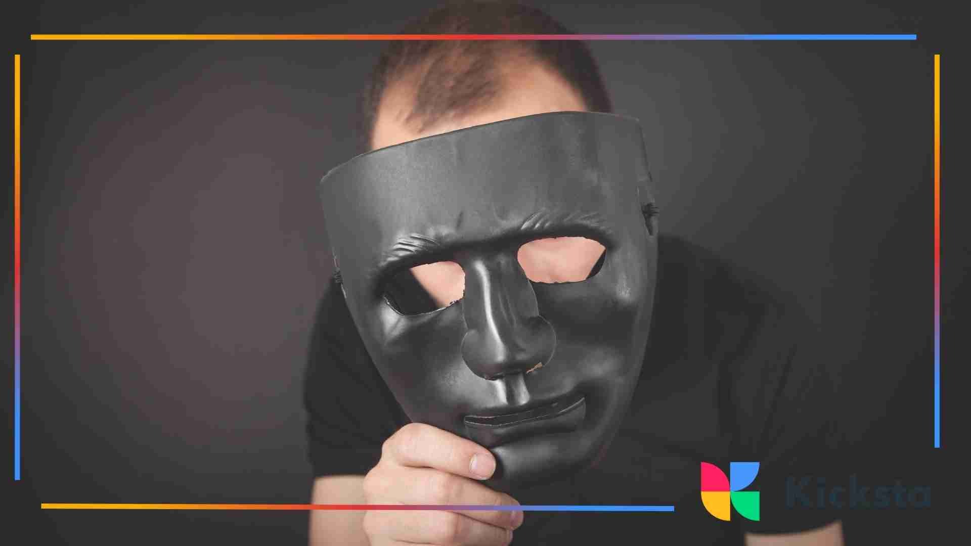 Person holding a black mask in front of their face against a dark background, representing anonymity or hidden identity.