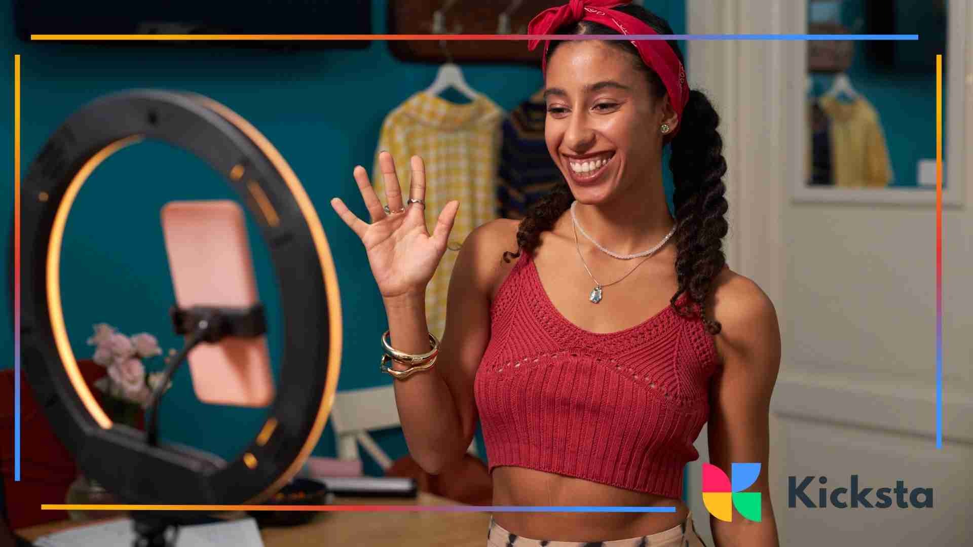 Woman smiling and waving while recording a video on a smartphone with a ring light.