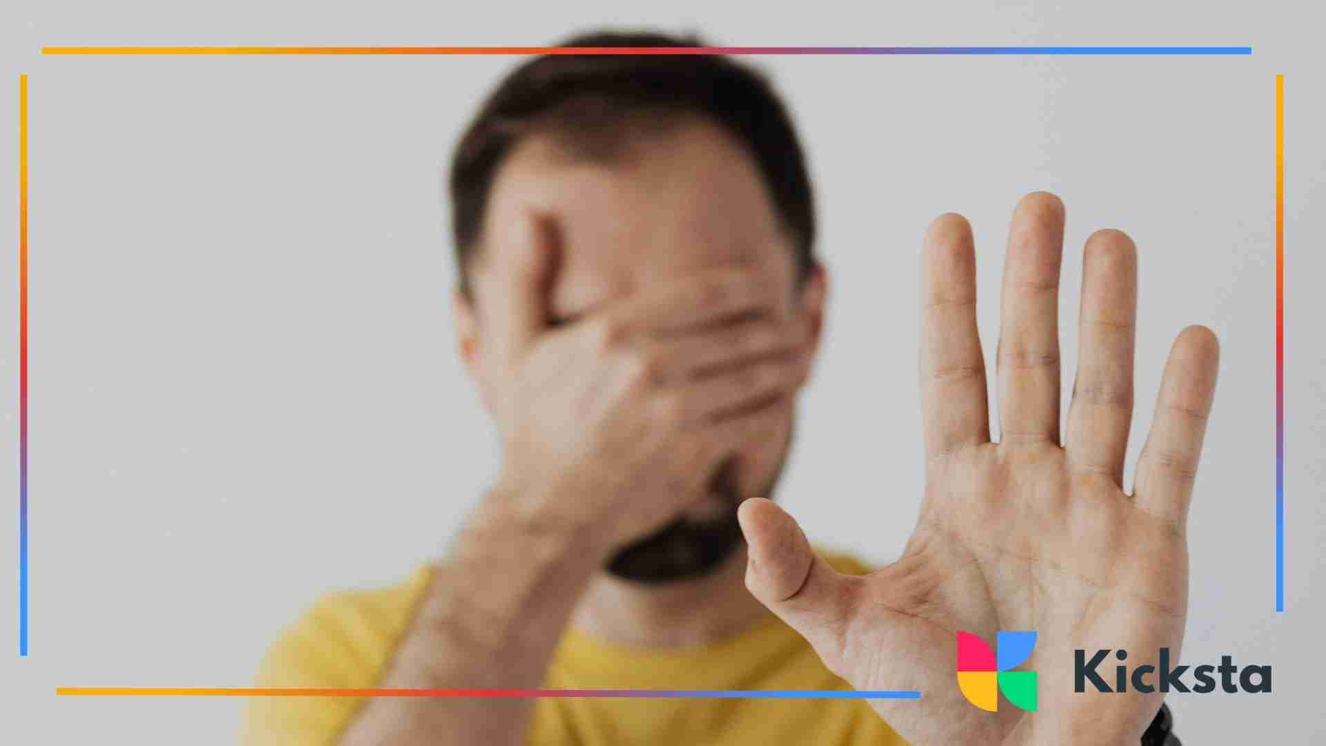 Person covering their face with one hand while holding the other hand up in a stop gesture.