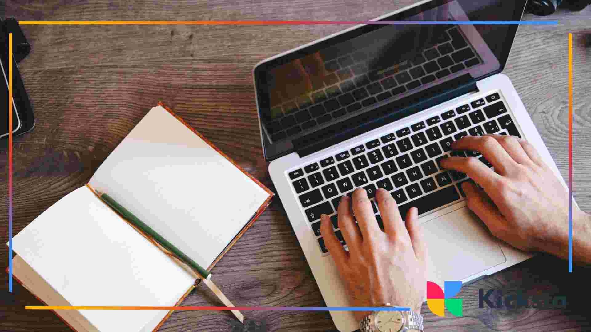 Hands typing on a laptop next to an open notebook and pencil on a desk.