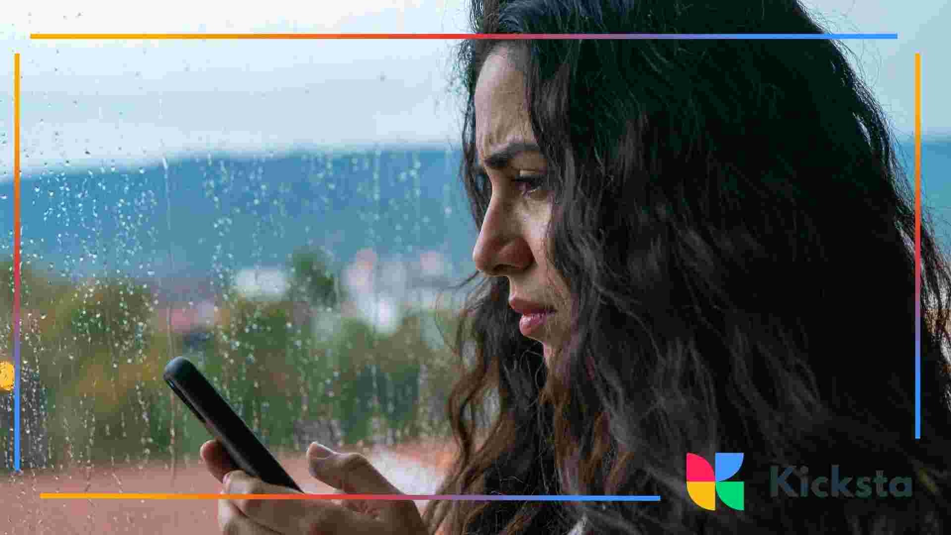 Person with long dark hair looking concerned while checking a smartphone near a rainy window.