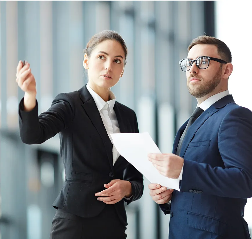 Two young business professionals in suits discussing paperwork, with the woman gesturing to the side.