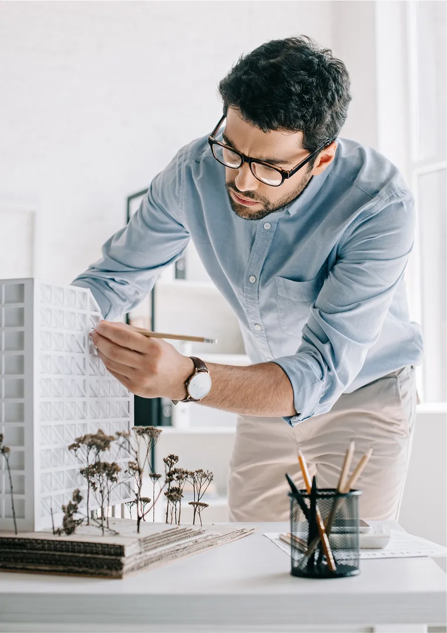Architect in glasses adjusting a white architectural model on a desk with dried plants and pencils.