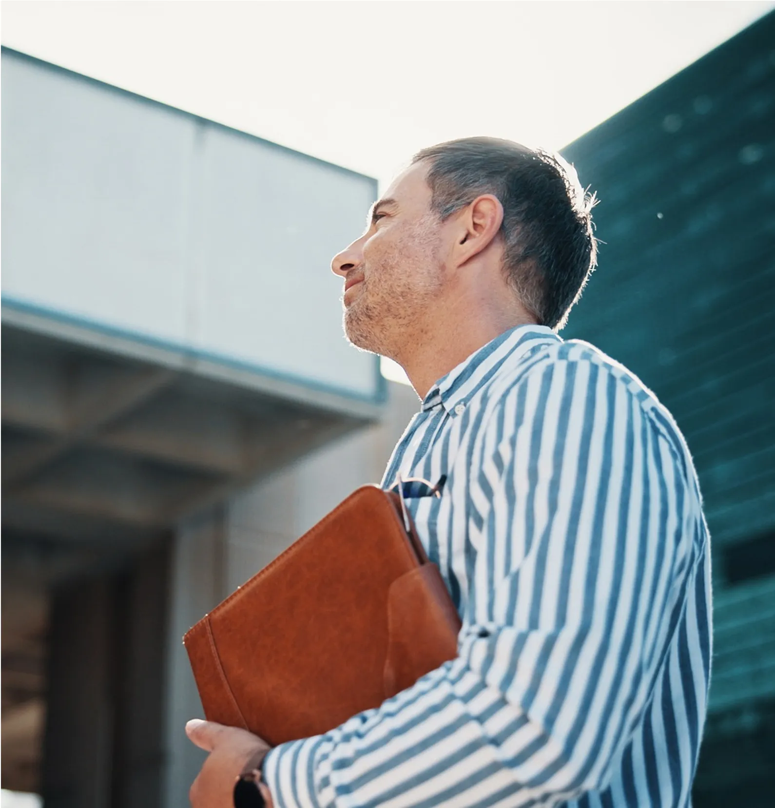 Man in a striped shirt holding a brown leather portfolio, standing outdoors near modern buildings.