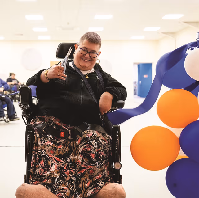 A service user (in a wheelchair) performs the ribbon cutting for the Paul's Place Hub, surrounded by blue and orange balloons and other celebrants.