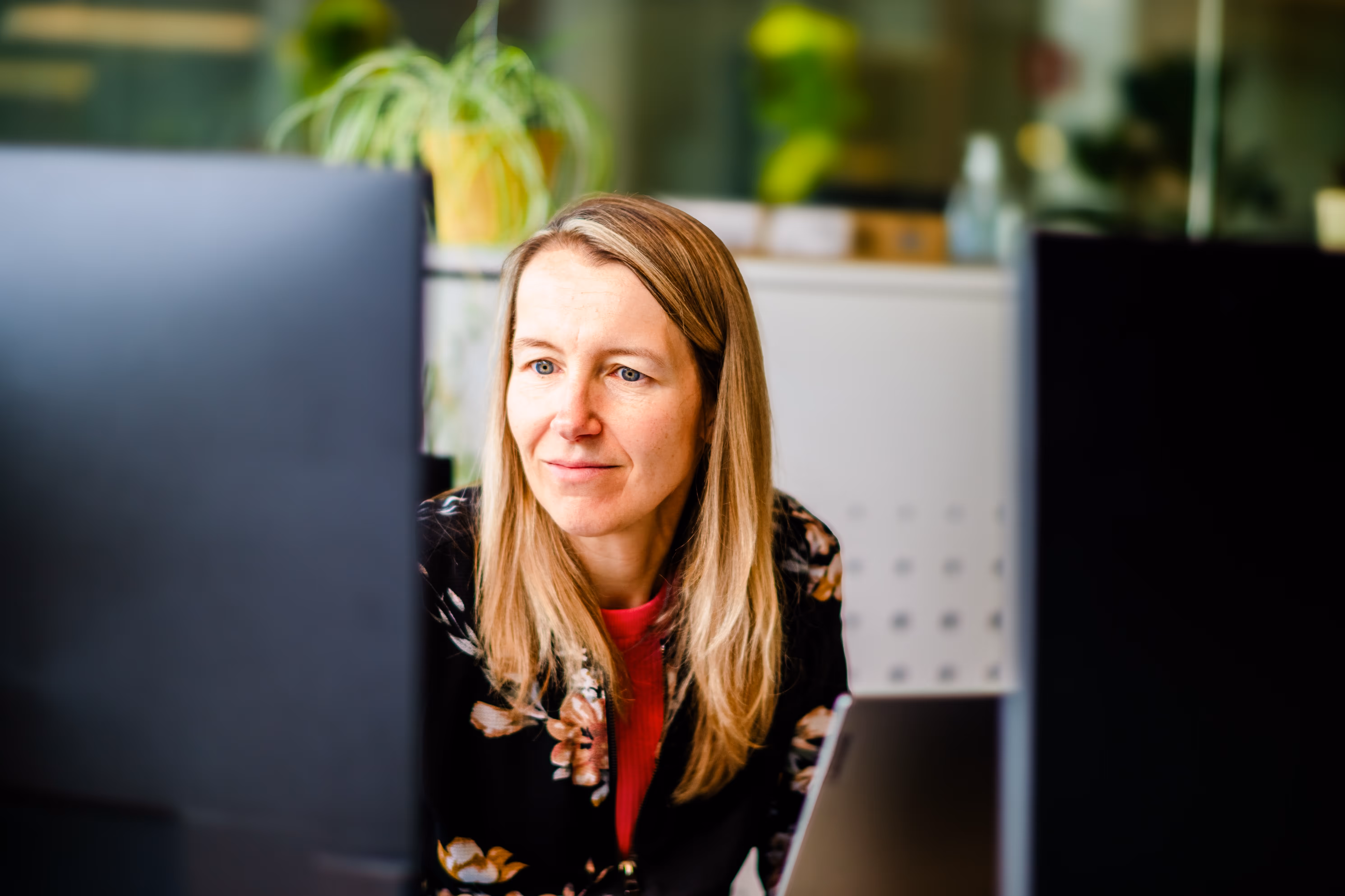 A woman focused on her computer screen, working in a bright office space with green plants in the background.