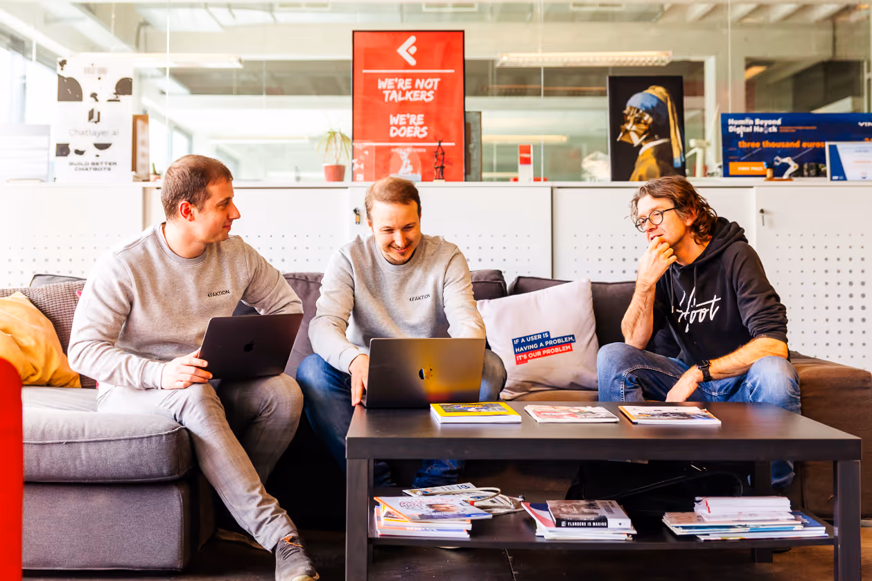 Three Faktion team members sitting on a couch, collaborating with laptops and smiling in a casual office setting.