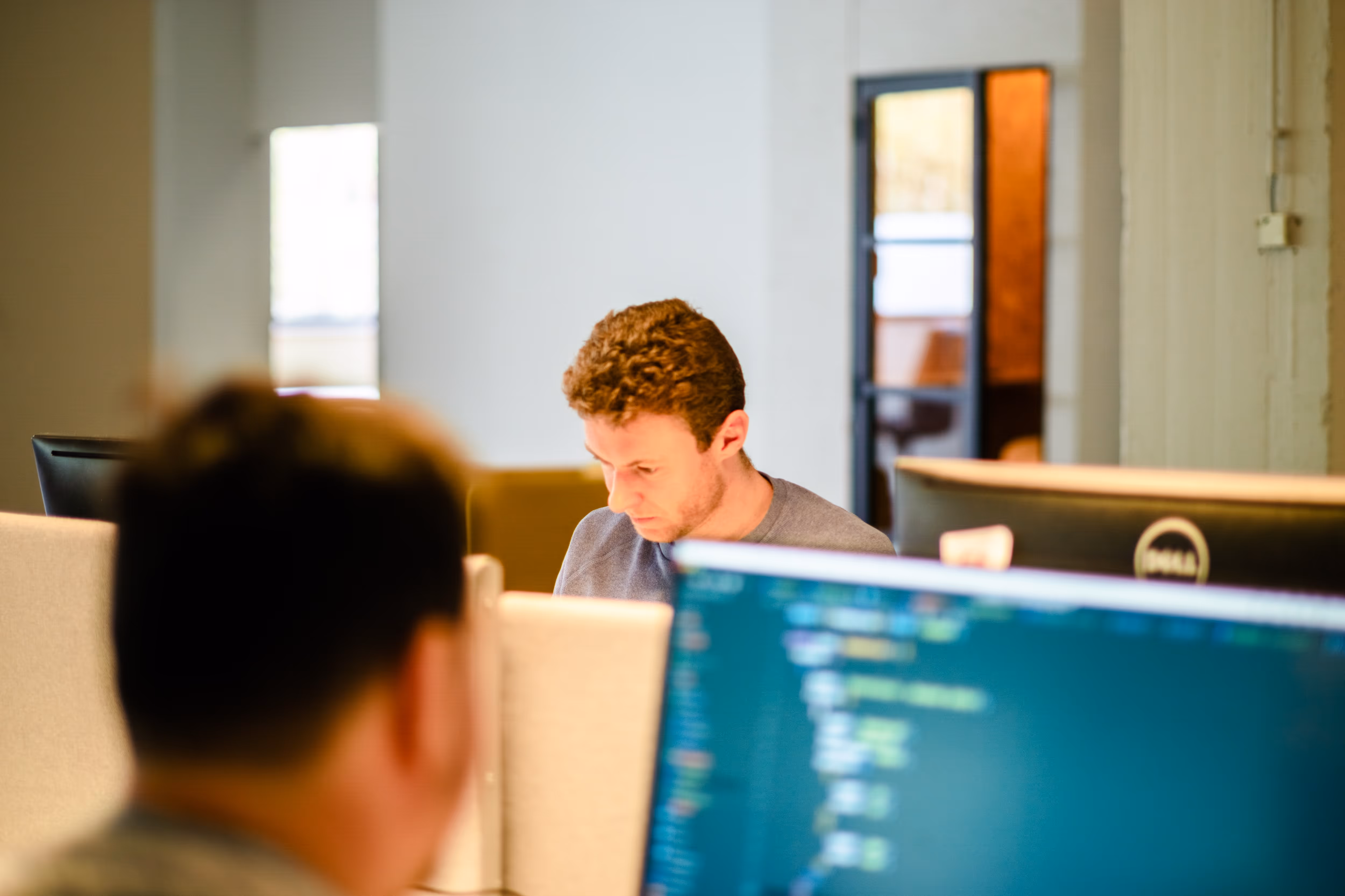 Two men working in an office with computer screens showing code, one man focused on his work in the background.
