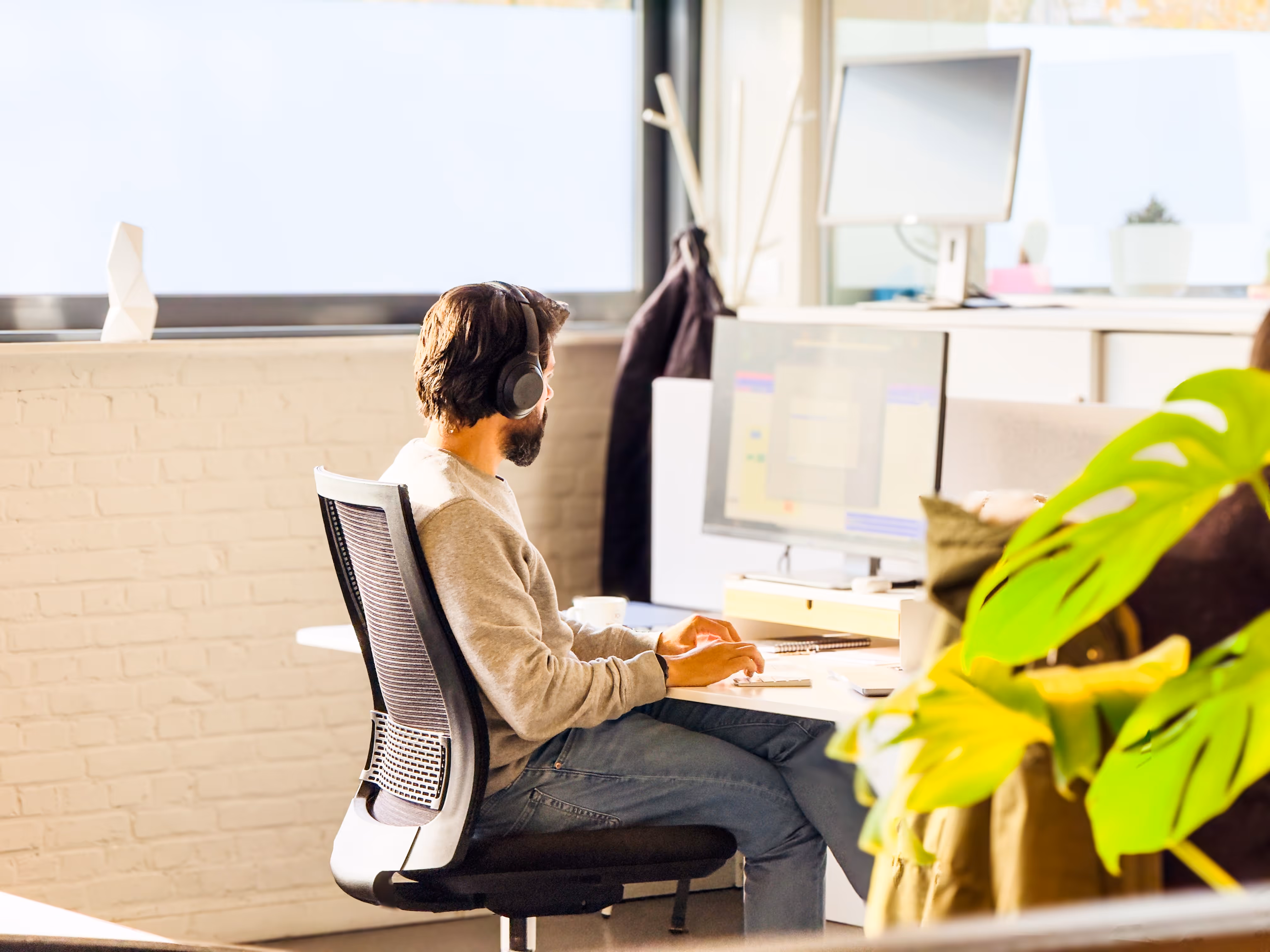 A Man wearing headphones is working on a computer at a desk in a bright office.