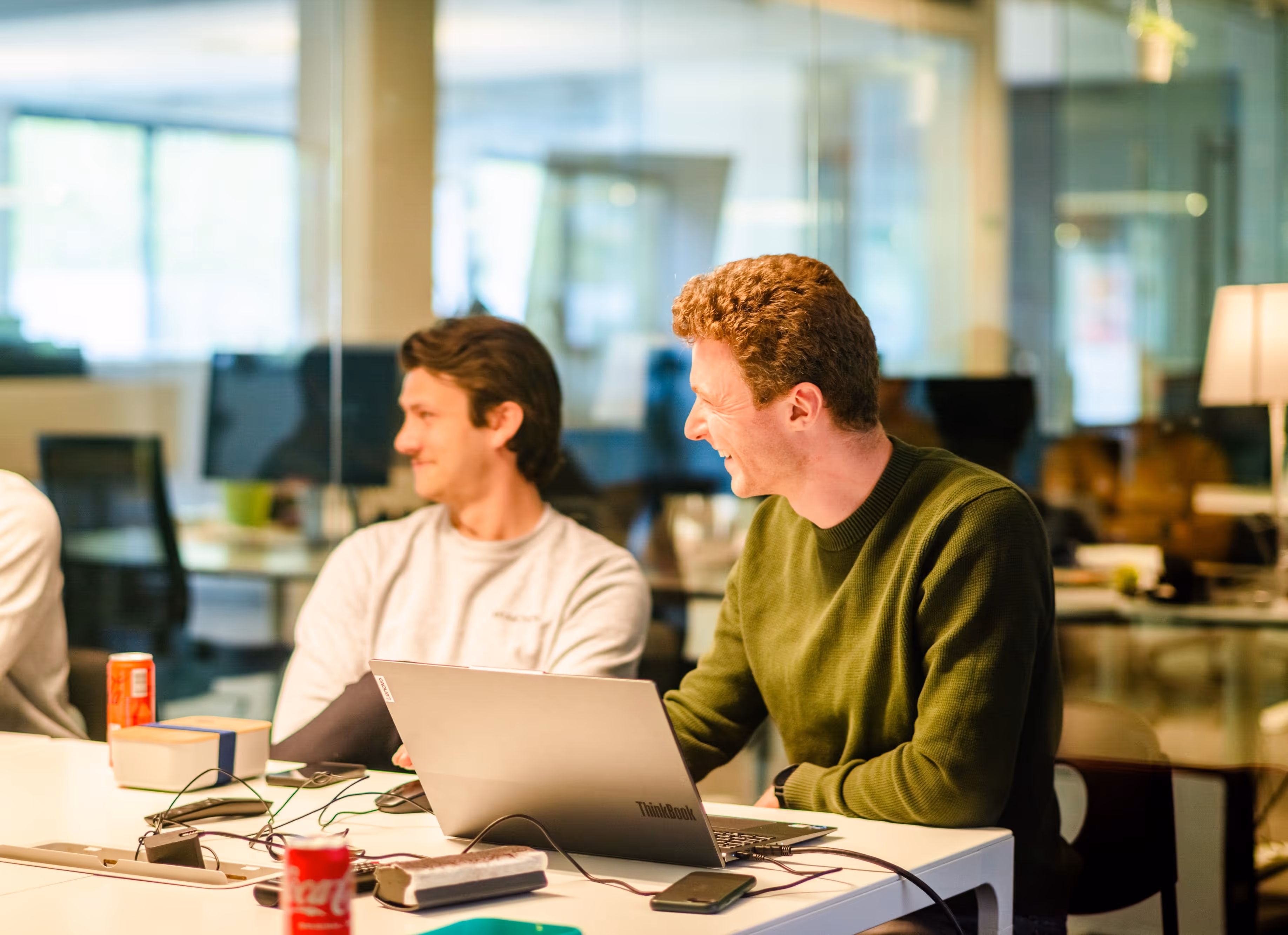 Two young men sitting at a table in an office, smiling and looking to the side with a laptop and soda cans on the table.