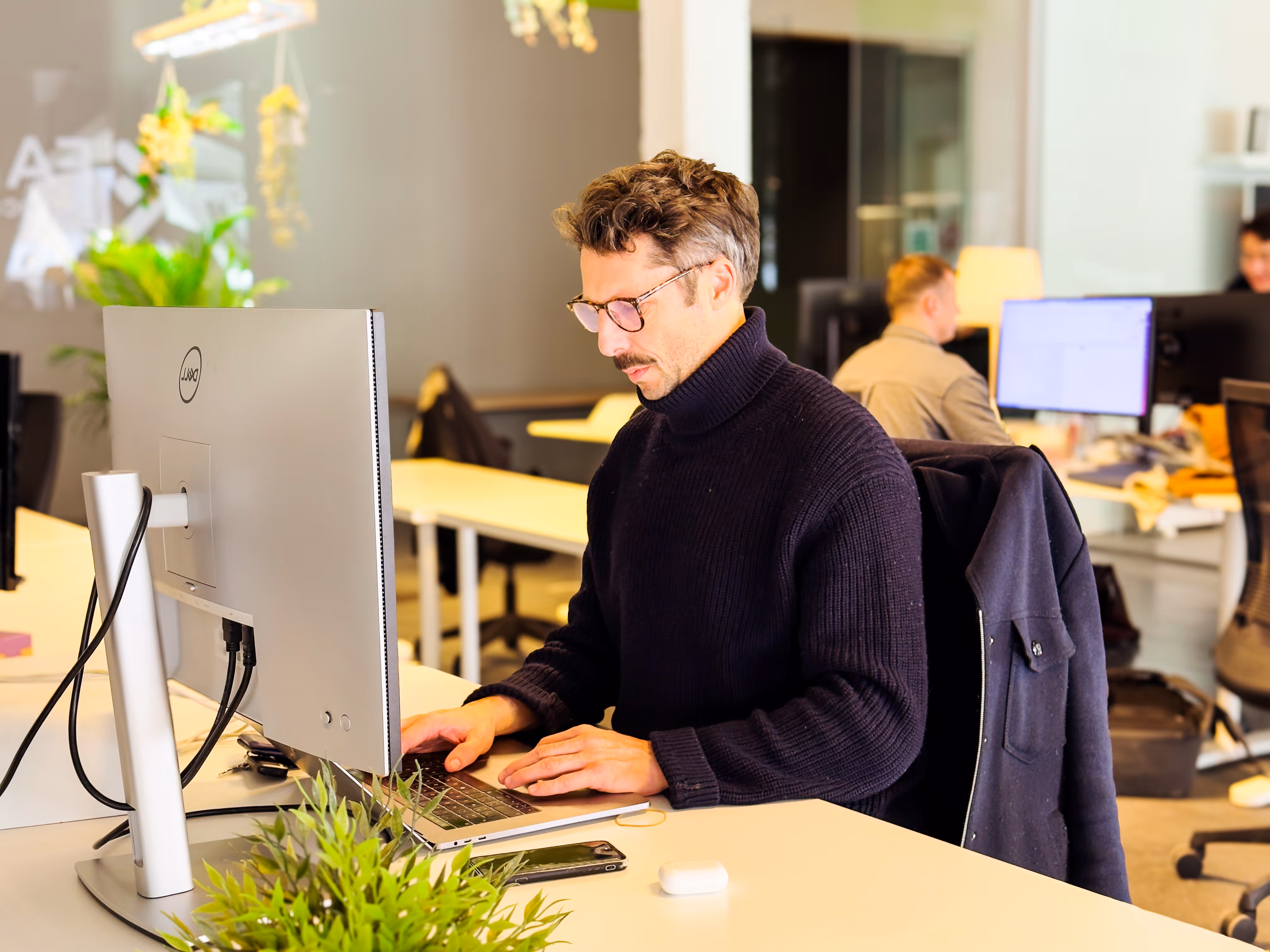 A man in glasses and a dark sweater working intently at a desktop computer in a modern open office.
