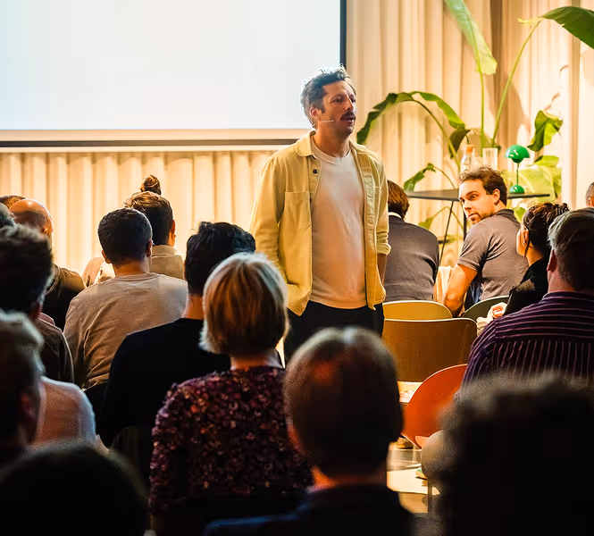 Jan is addressing an audience in a well-lit room with plants and a projection screen.