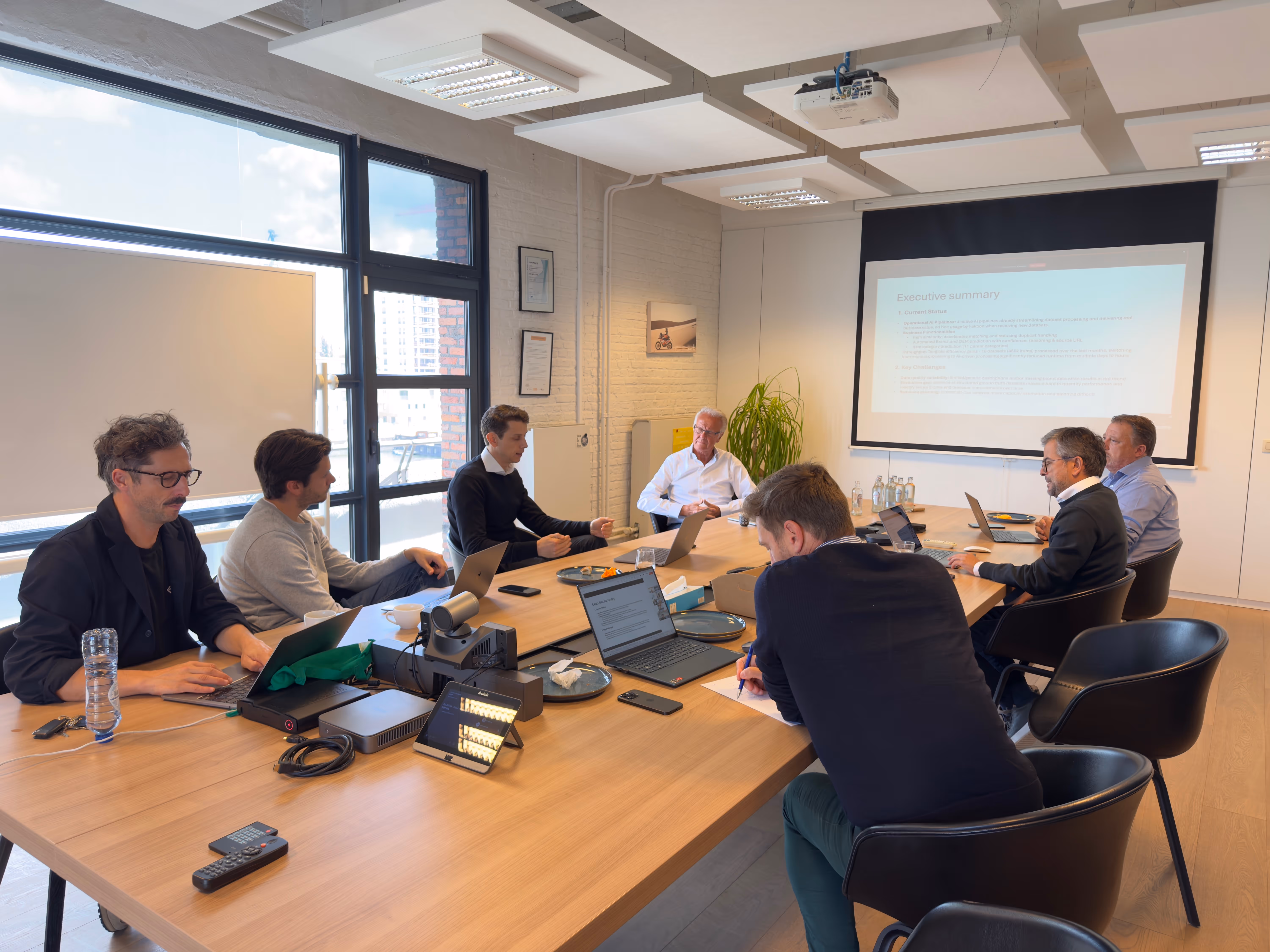 Seven people seated around a conference table with laptops, a projector screen showing a presentation, in a bright meeting room with large windows.